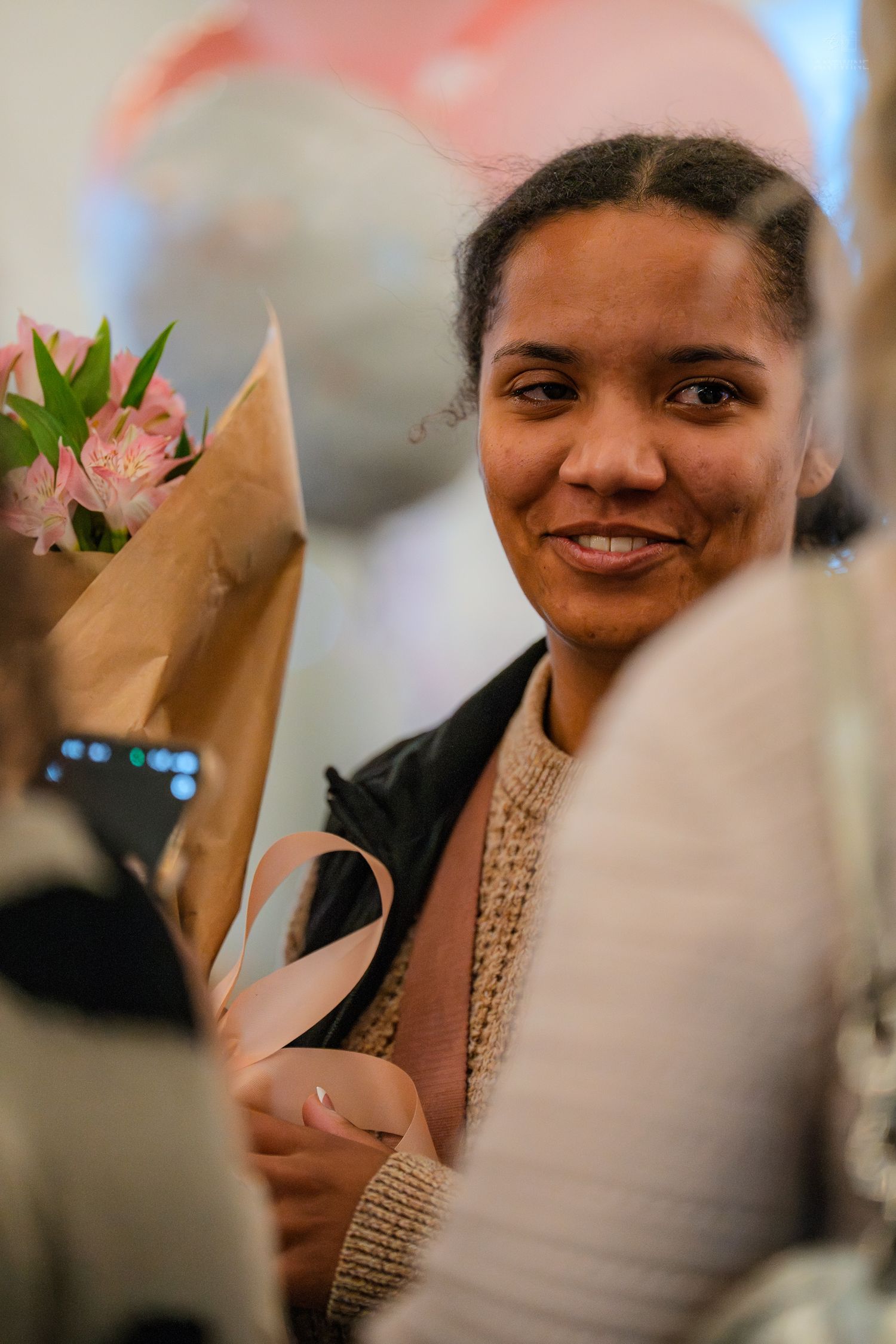 A young woman smiles holding a pink flower bouquet wrapped in kraft paper at a joyful celebration.