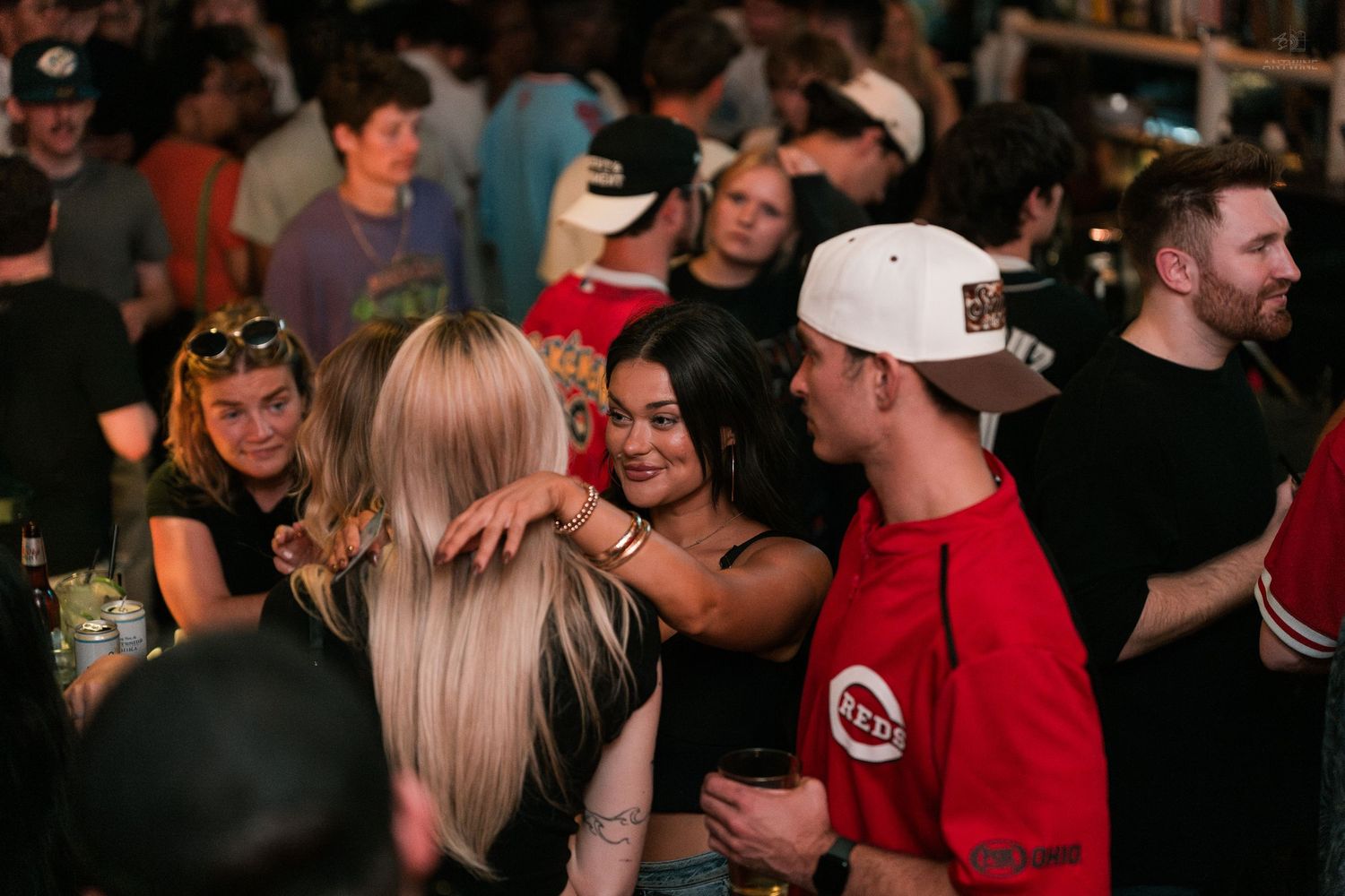 A group of young adults hugging and laughing together in a crowded bar with warm lighting.