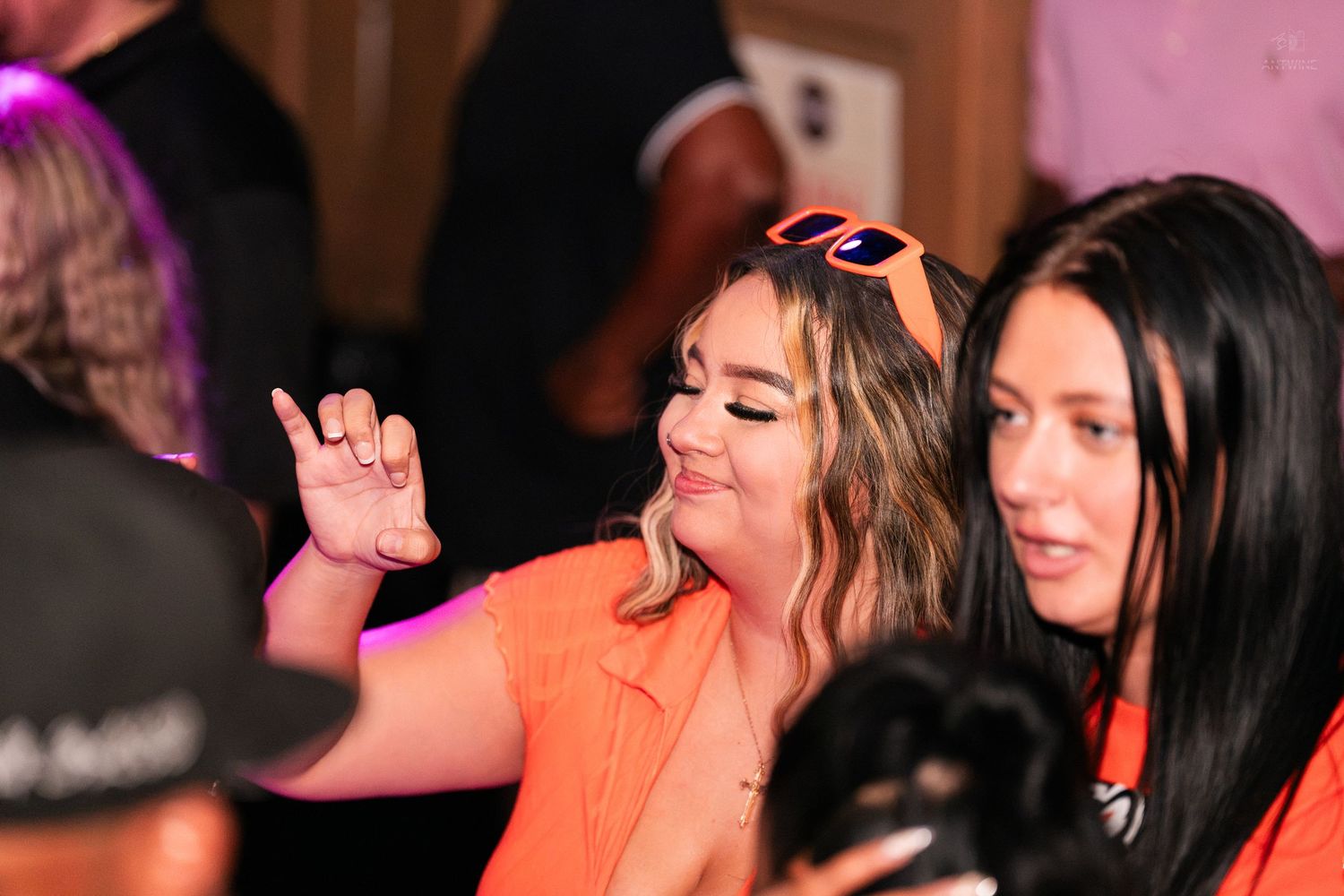 A woman in orange dances with hand gestures beside a friend at a lively party.