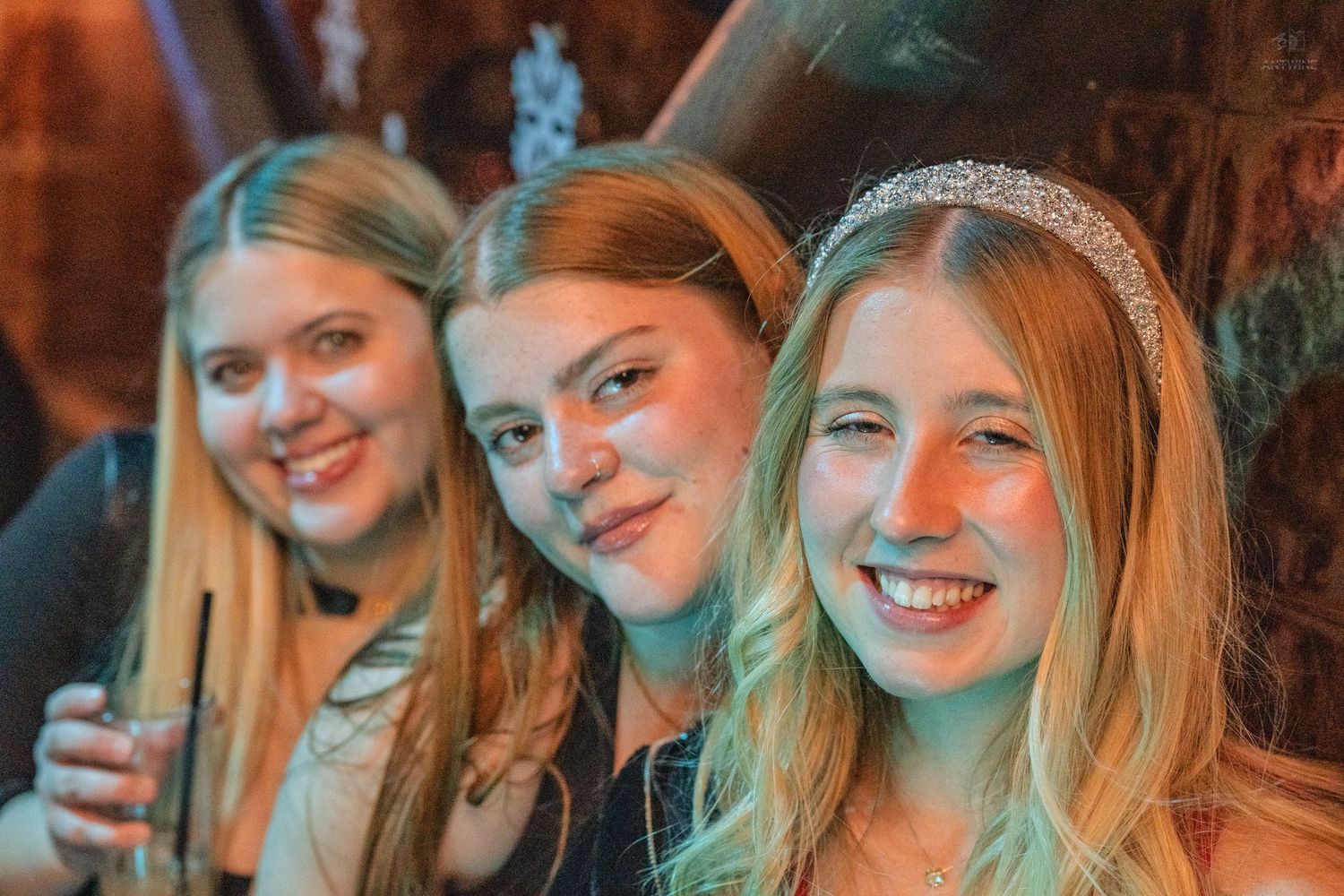Three young women smiling at a party, one holding a drink and wearing a rhinestone headband.
