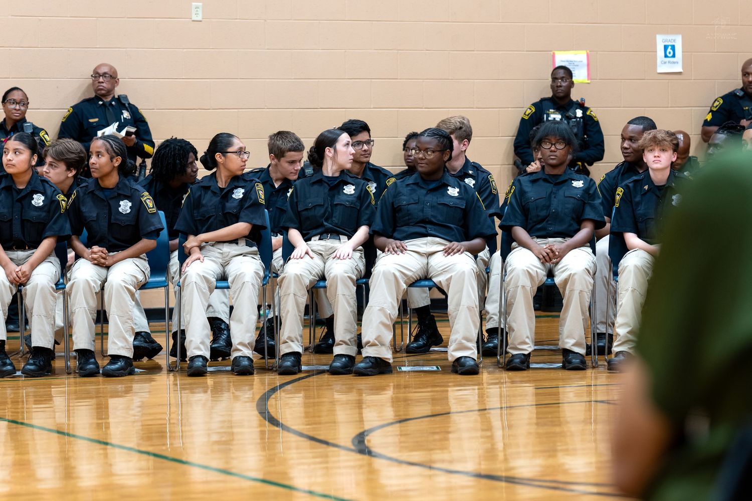 Students in uniform sit in rows of chairs inside a school gymnasium during a formal law enforcement youth program ceremony.