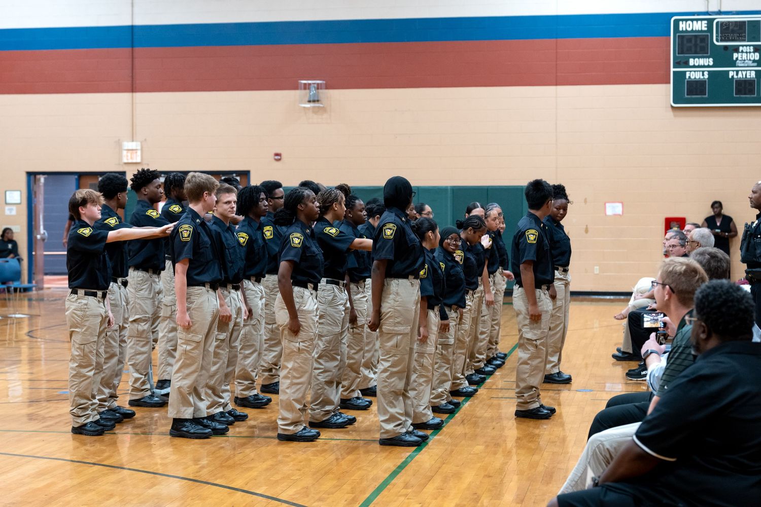Students in uniform khaki and navy shirts stand in formation on a gymnasium floor during a school JROTC ceremony.