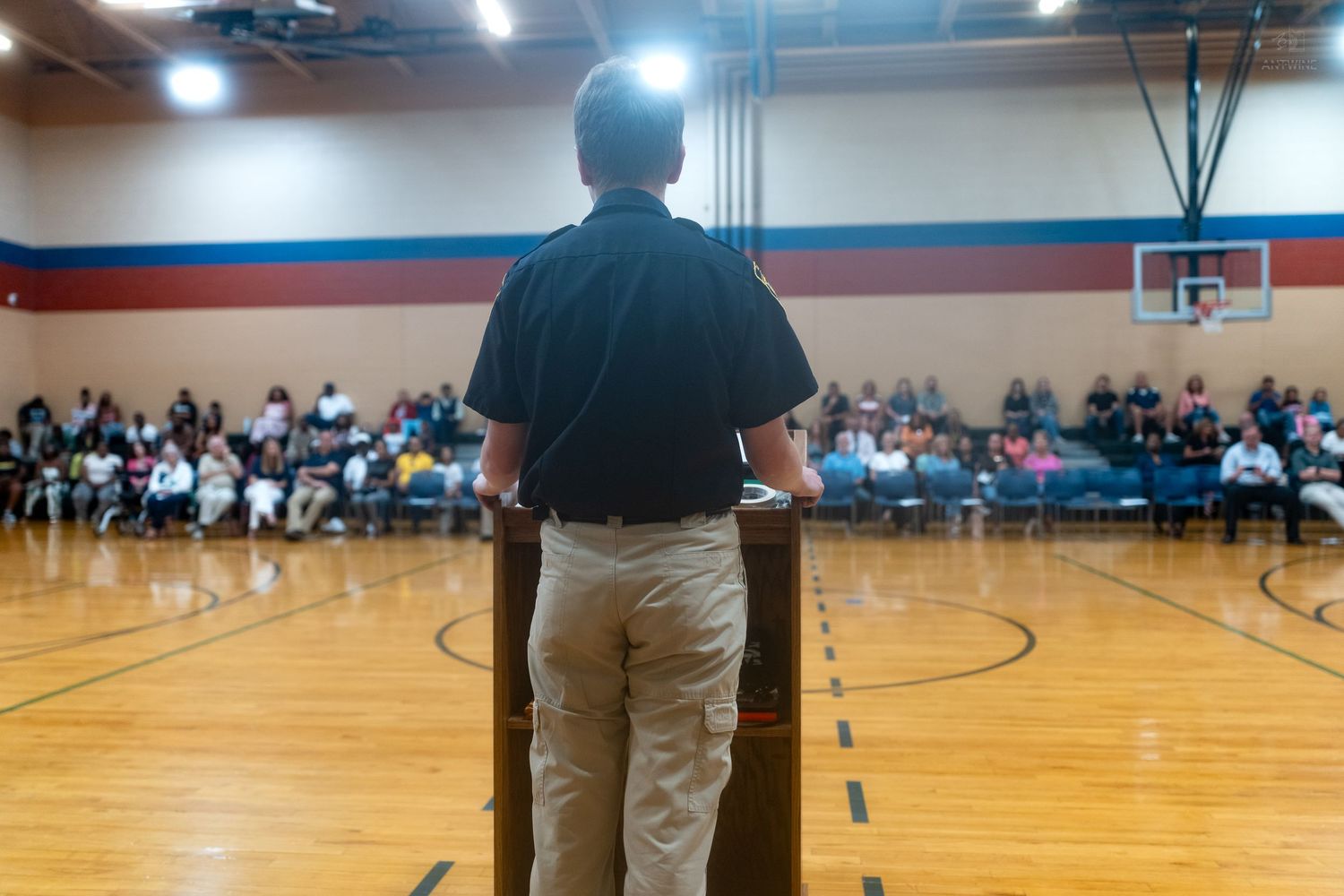 Person speaking at podium in school gymnasium to large seated audience during assembly event.