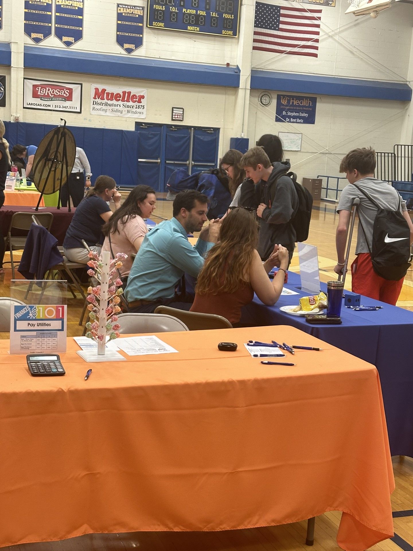 Students and vendors interact at tables during a school career fair in a gymnasium with blue and gold decor.