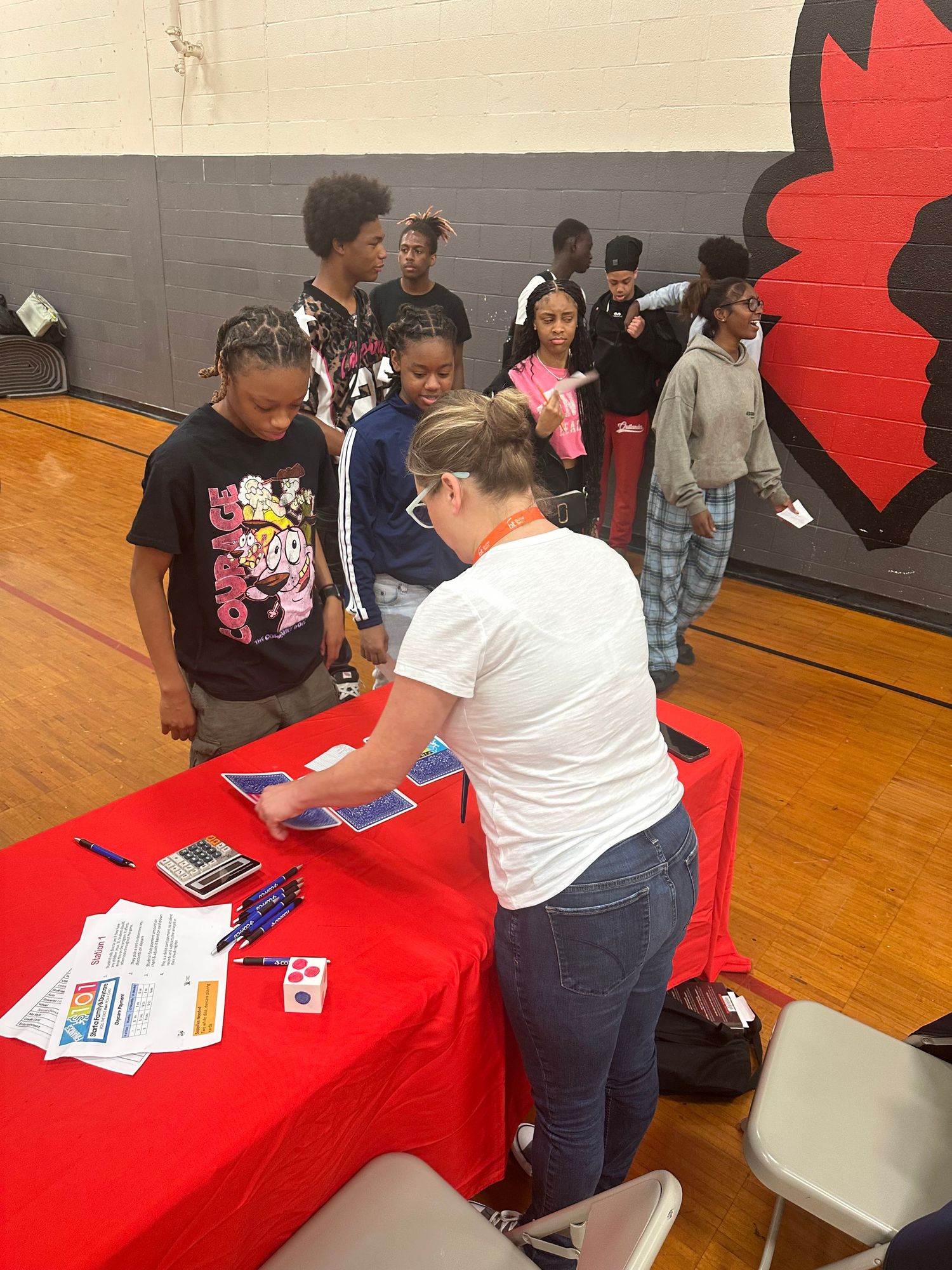 A group of students lines up at a red table as a representative hands out materials at a school career fair.
