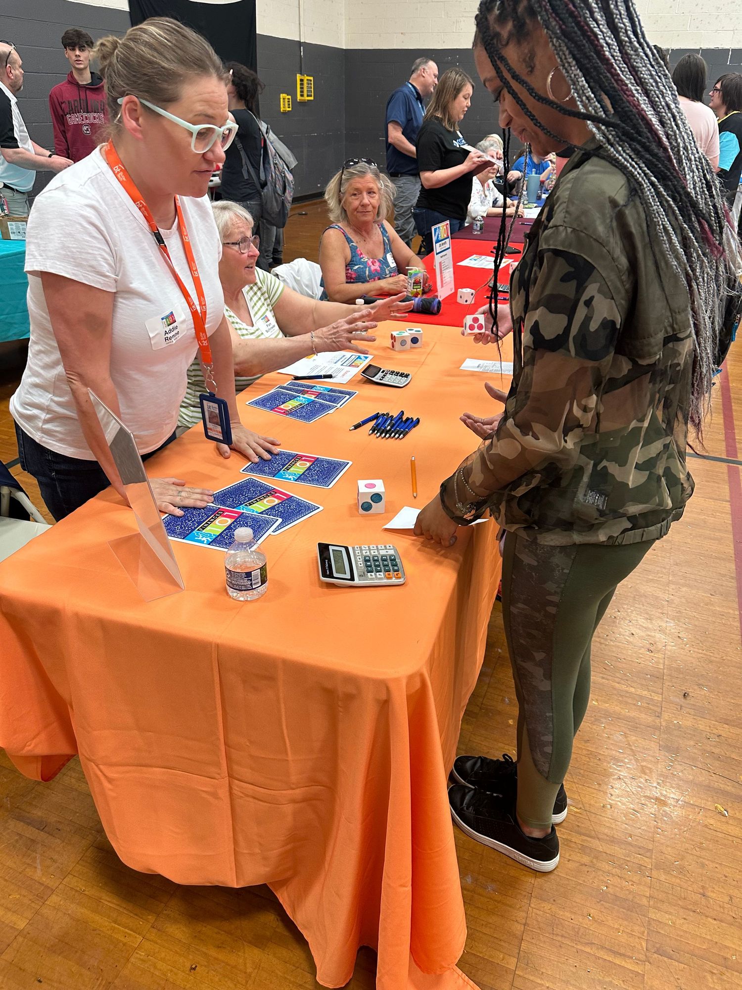 Female presenter shows colorful educational materials at an orange table to a student at an indoor school gymnasium fair.