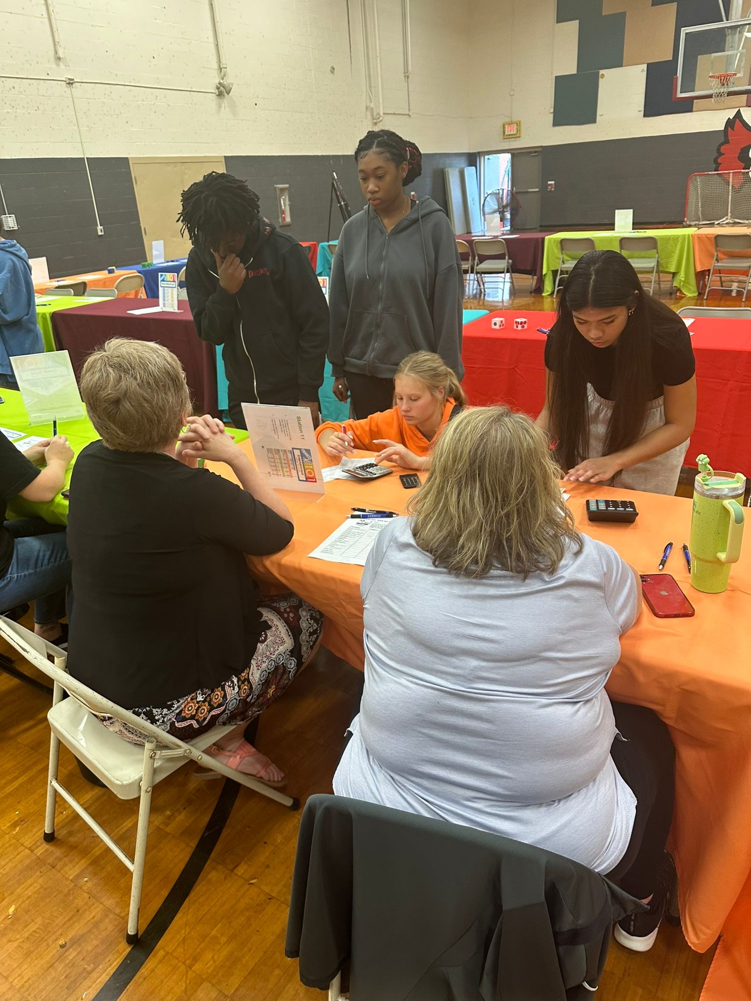 Students and adults gather around an orange table during a school career fair event in a gymnasium.