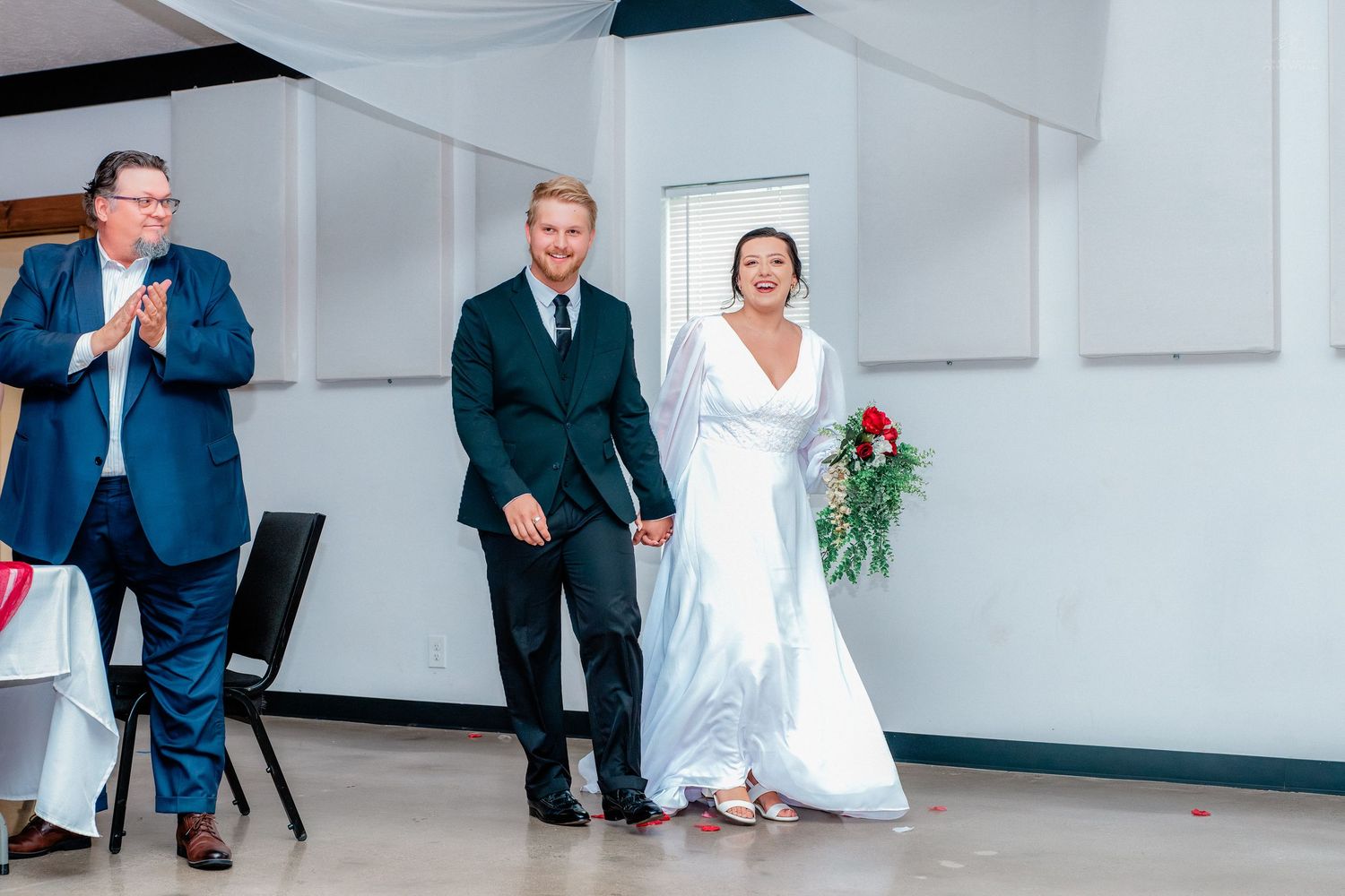 Smiling bride in white gown holding red roses walks with groom in dark suit at wedding reception ceremony.