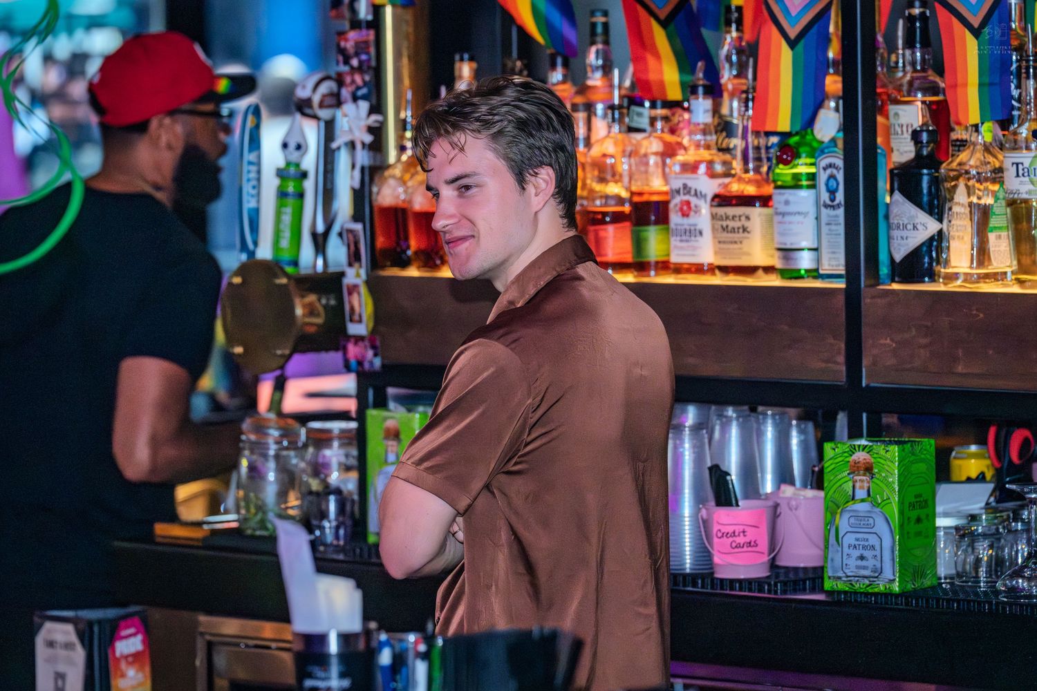 Young man in brown polo shirt standing at a colorful bar with liquor bottles and rainbow flag decor.
