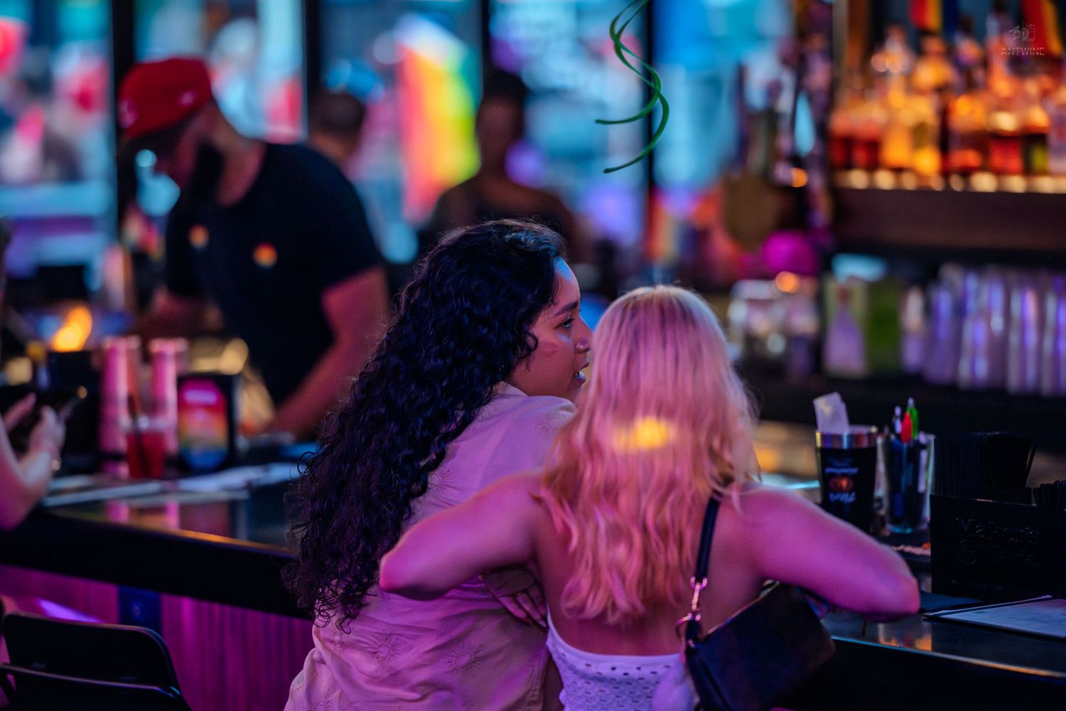Two women, one with dark hair and one with pink hair, embrace at a colorful neon-lit bar.