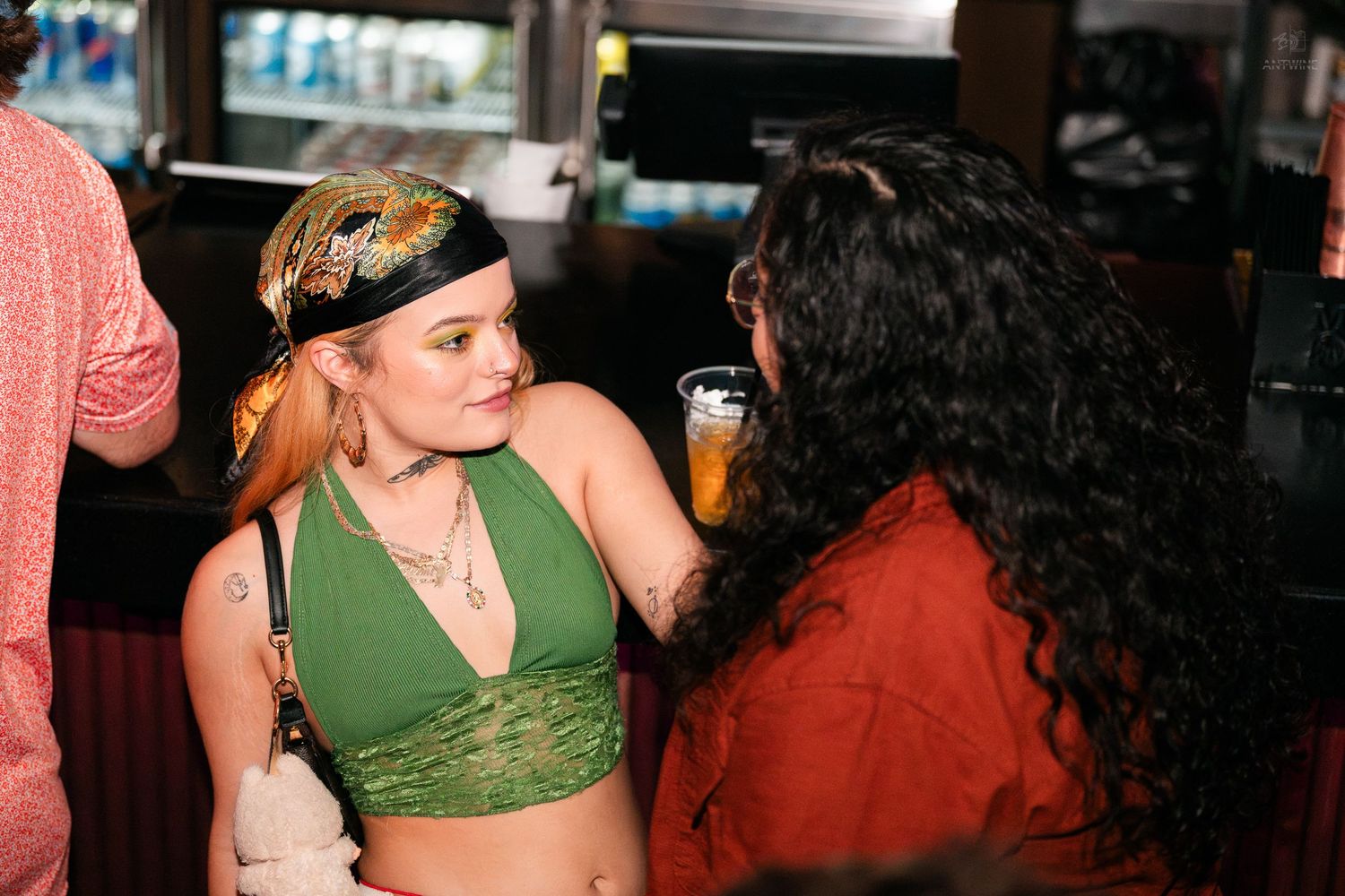 Two women chatting at a bar, one in a green crop top holding a drink.