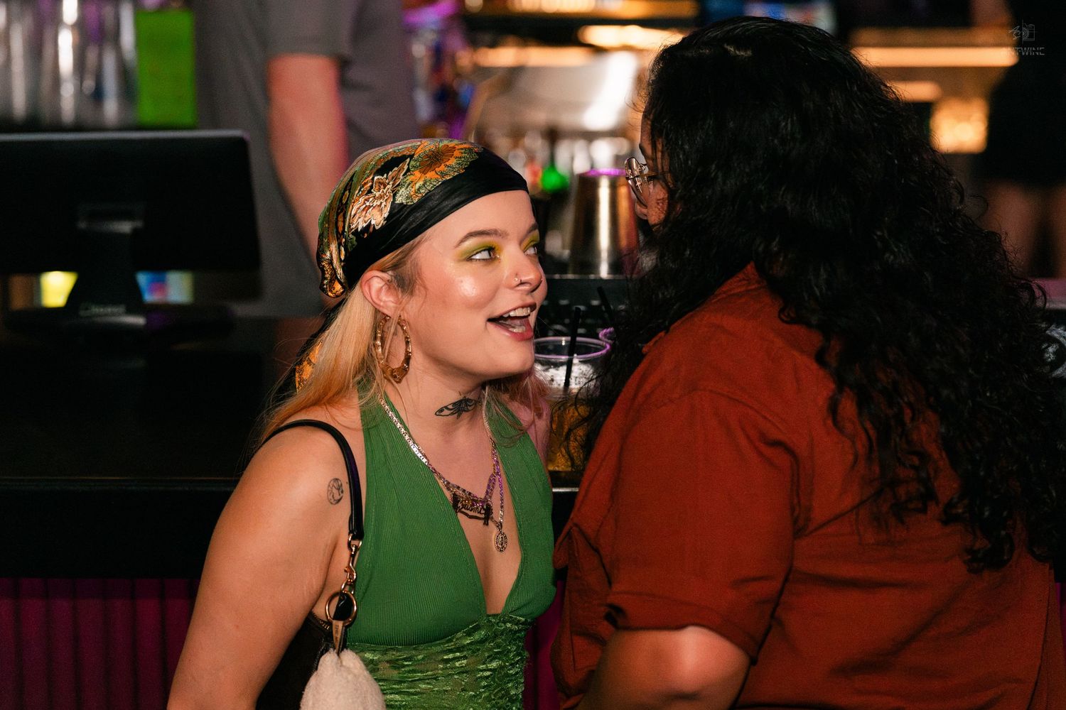 Two women laugh and chat together at a vibrant nightclub bar.