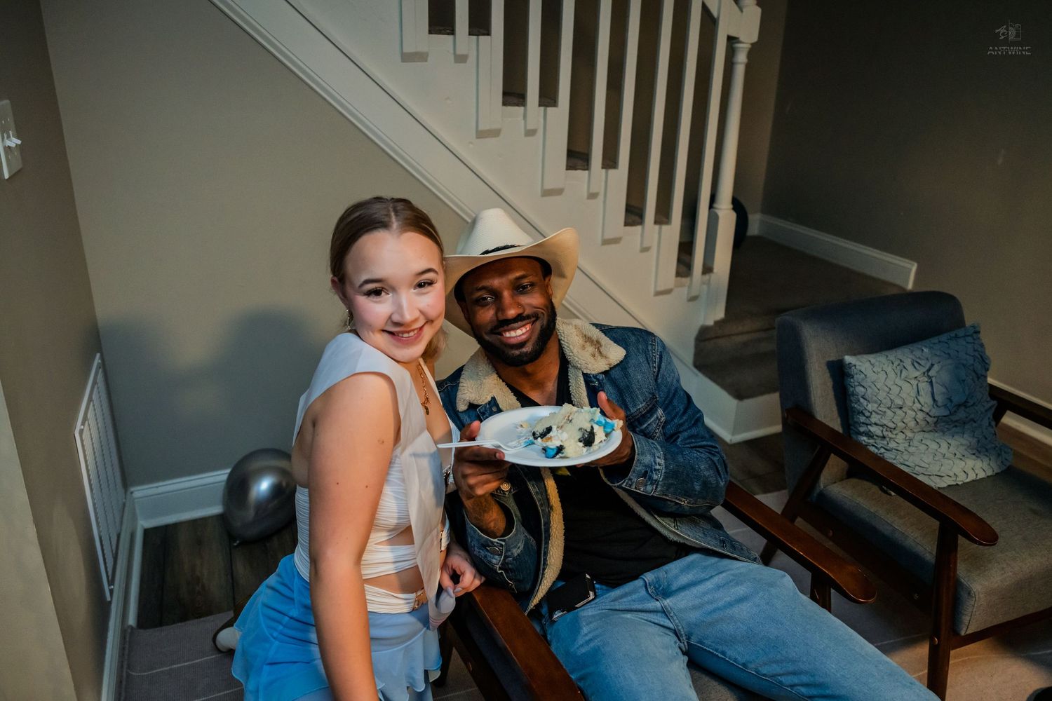 Young woman and man in cowboy hat pose together on stairs holding a plate of food at a birthday party.