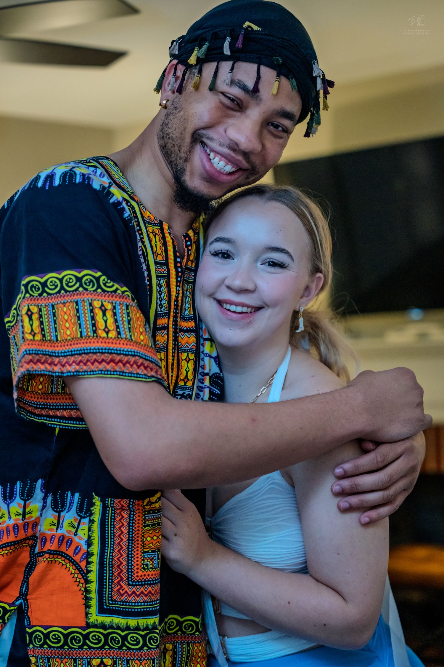 A cheerful young man in a vibrant dashiki shirt embraces a smiling young girl in a white outfit.
