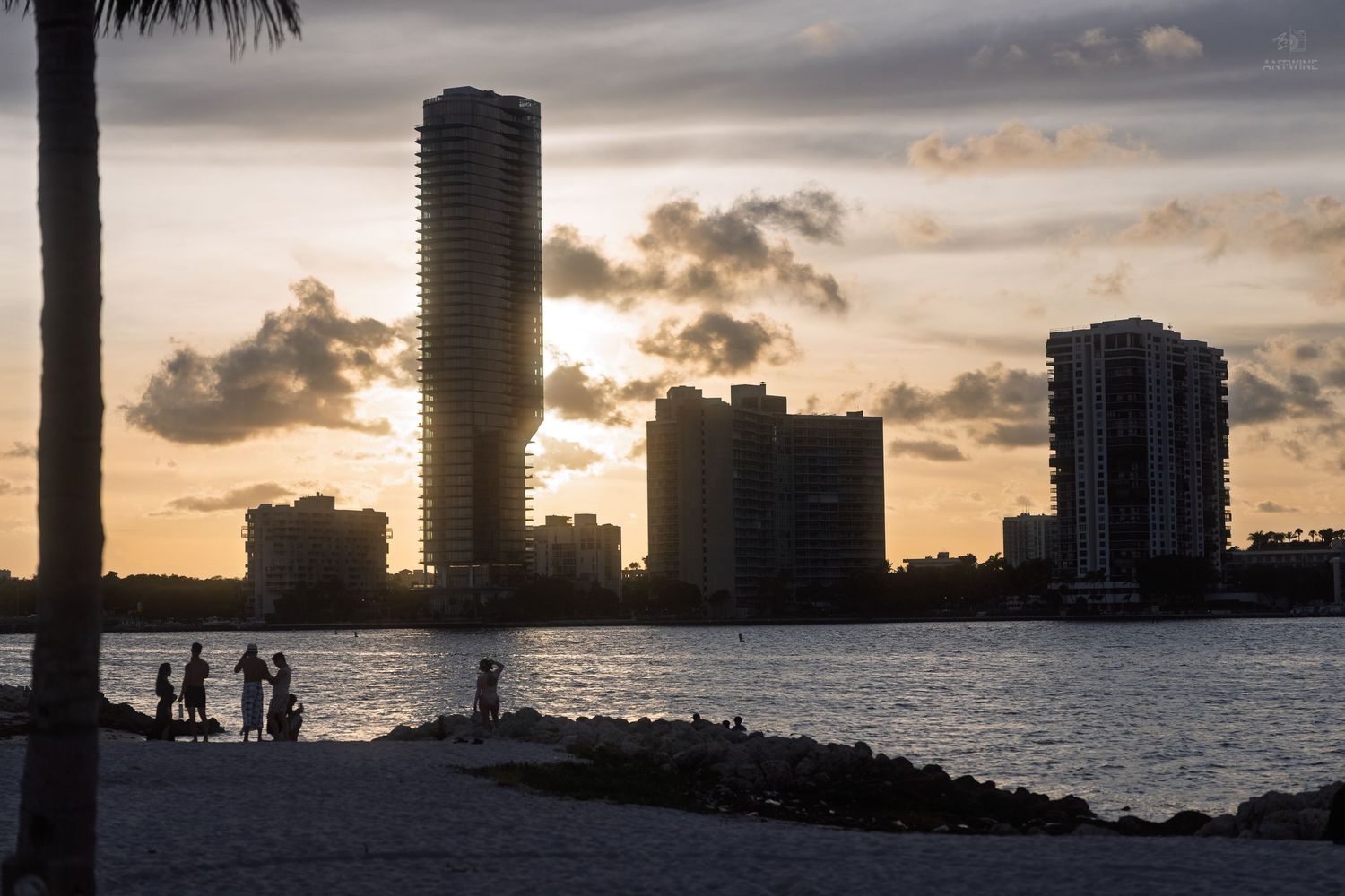 Silhouette of people on a sandy beach at sunset with tall Miami city skyscrapers reflected on calm water.