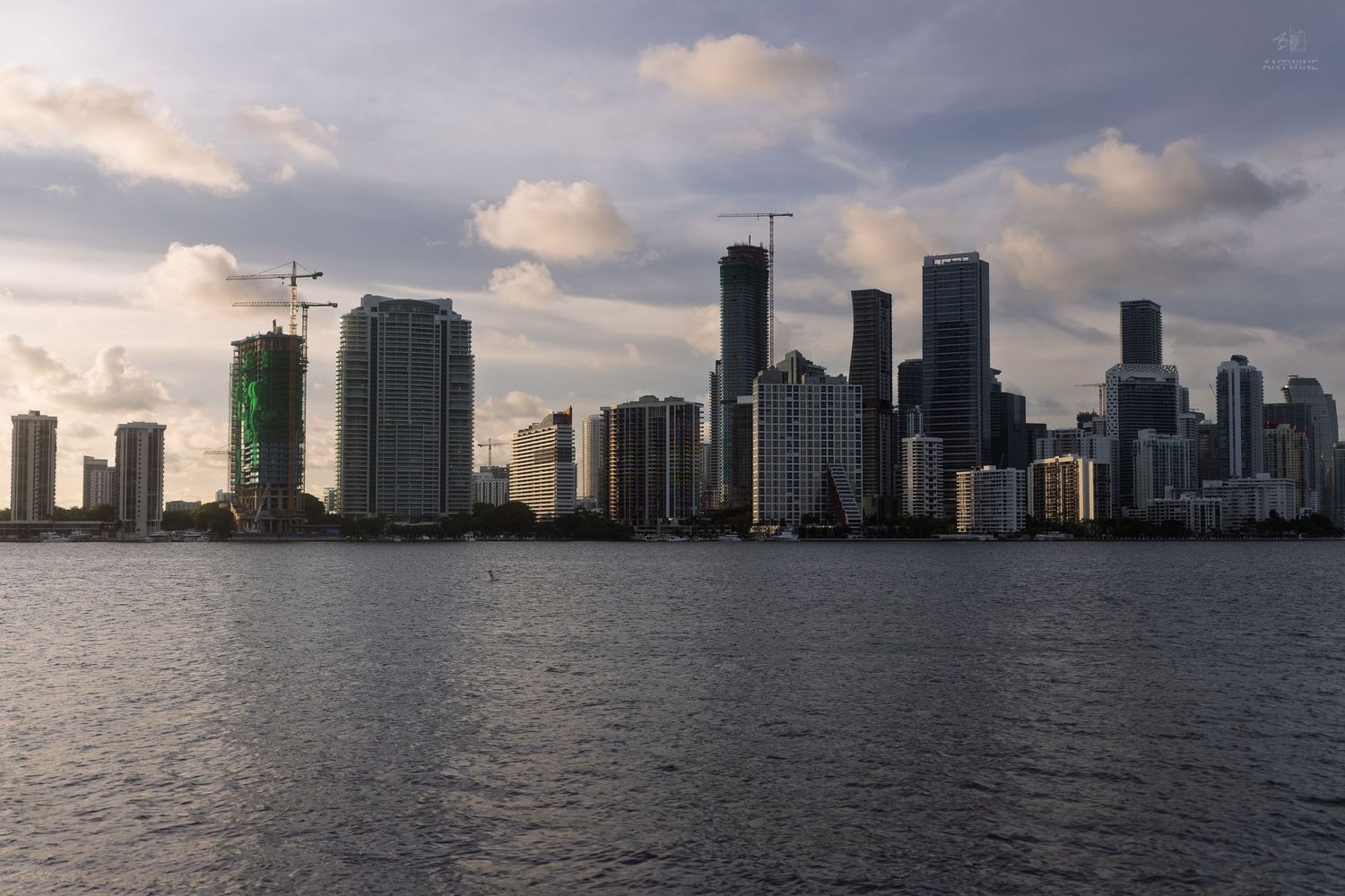 Miami downtown skyline with construction cranes reflected over Biscayne Bay at dramatic sunset.