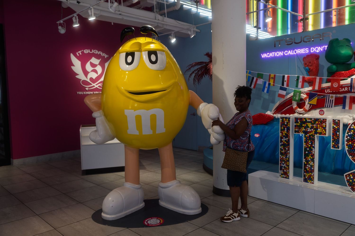 Child poses next to a large yellow M&M character statue inside a colorful candy store display.
