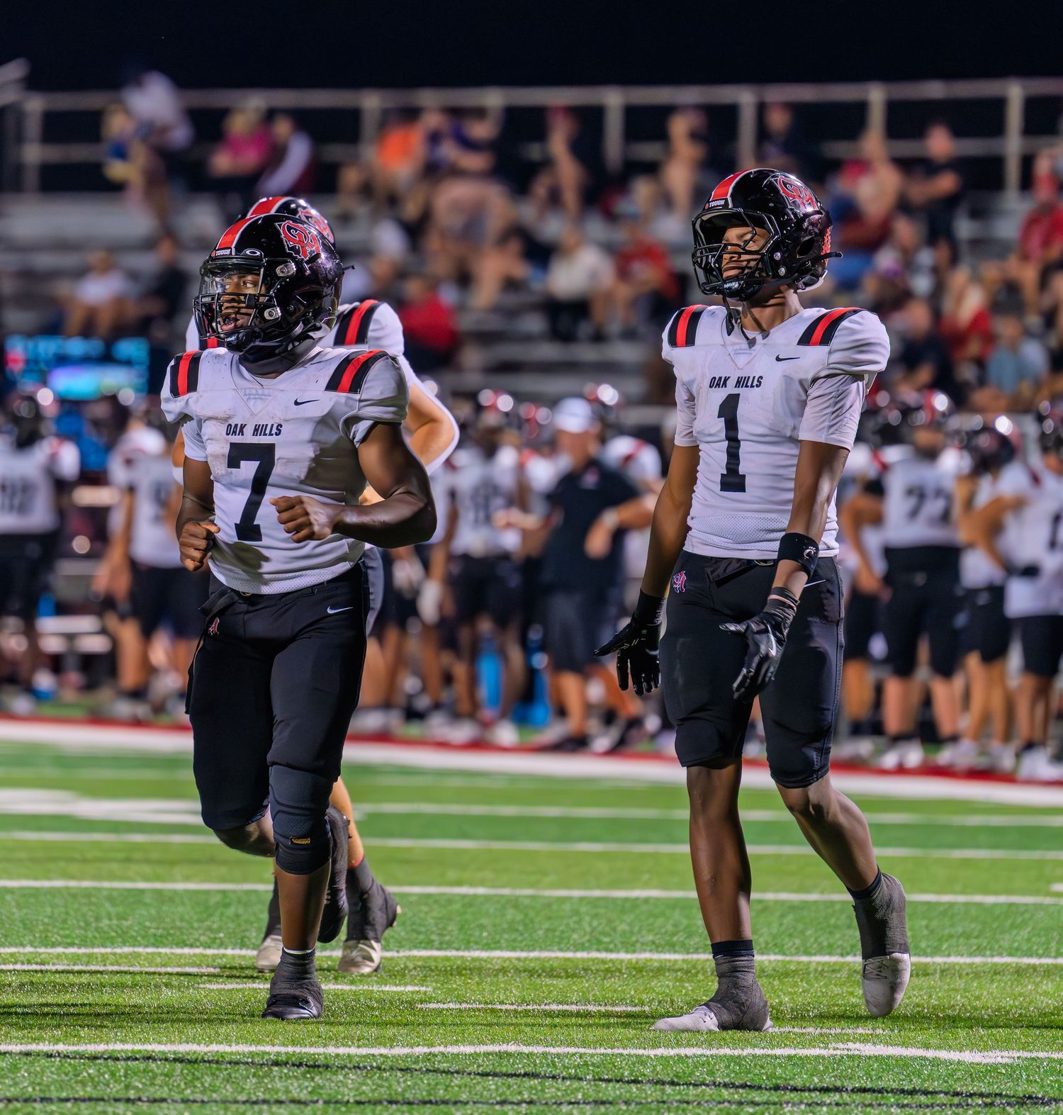 Two high school football players in red and black uniforms jog on the field during a night game.
