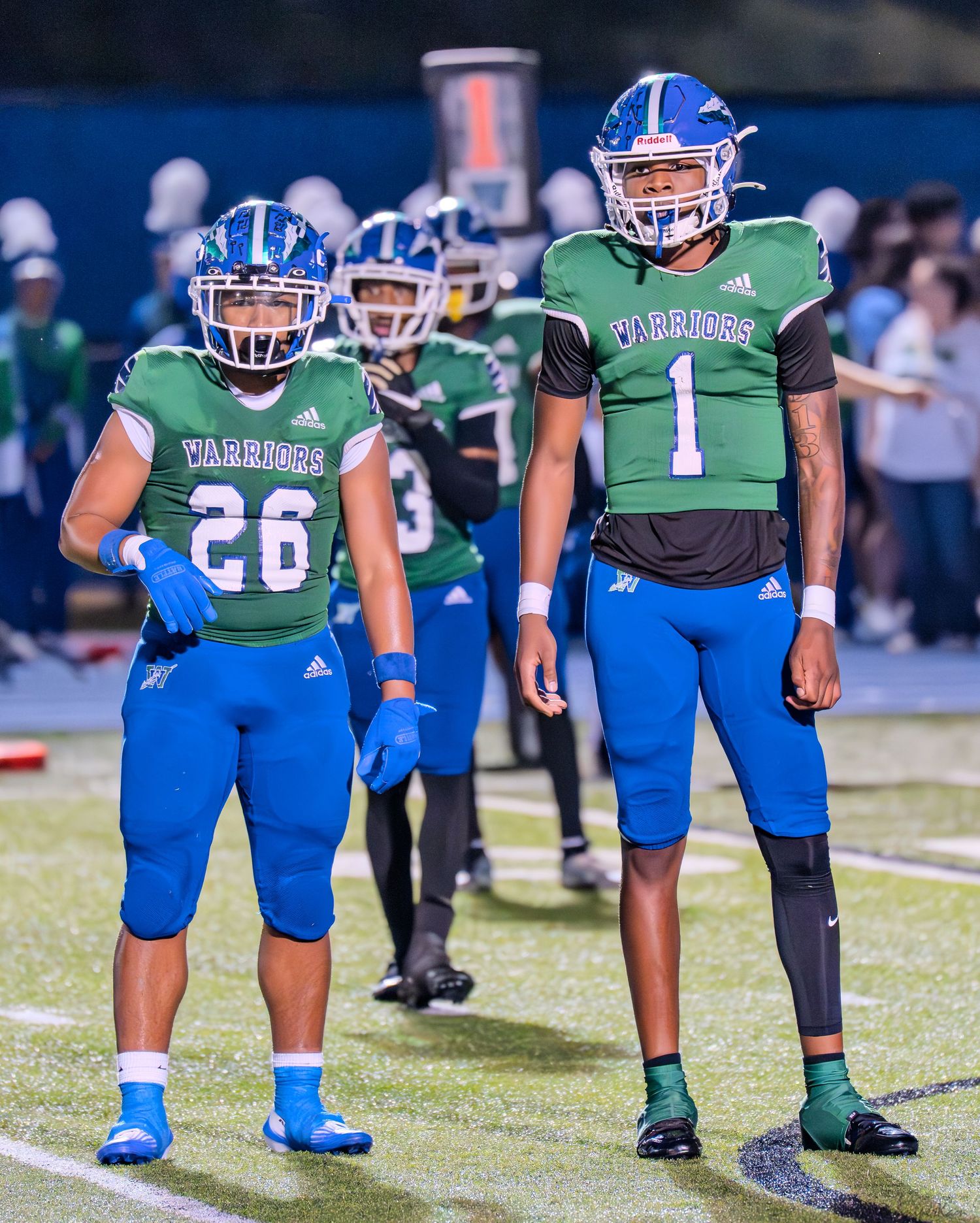 Two Warriors football players numbers 26 and 1 in green and blue uniforms stand on the field during a game.