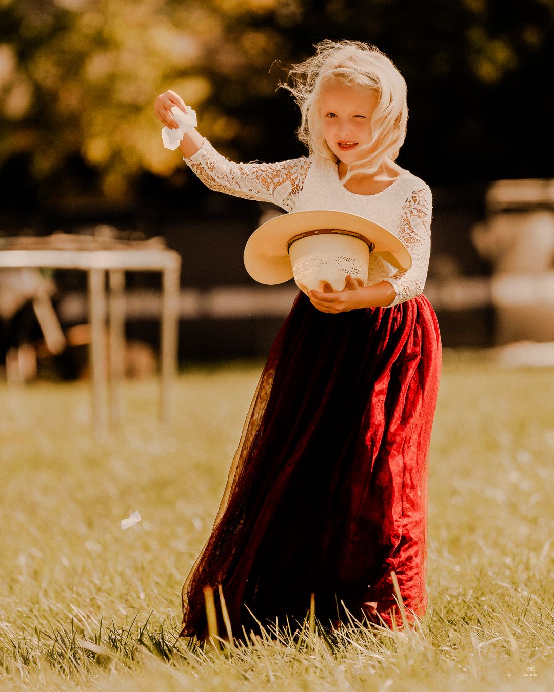 Young flower girl in white lace top and red skirt holding a hat while tossing petals outdoors.