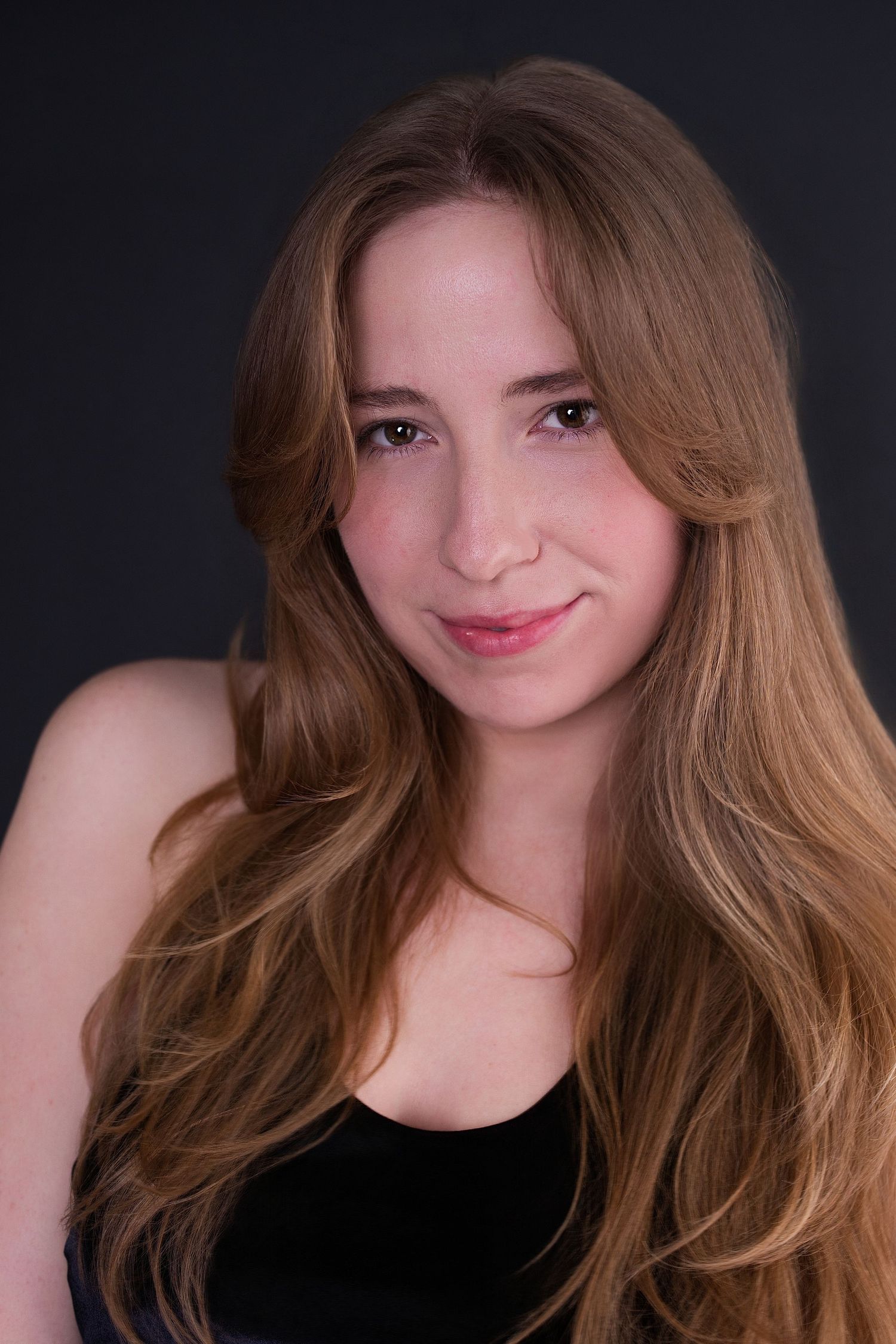 Young woman with long wavy blonde hair wearing a black top smiles for a professional headshot against a gray background.