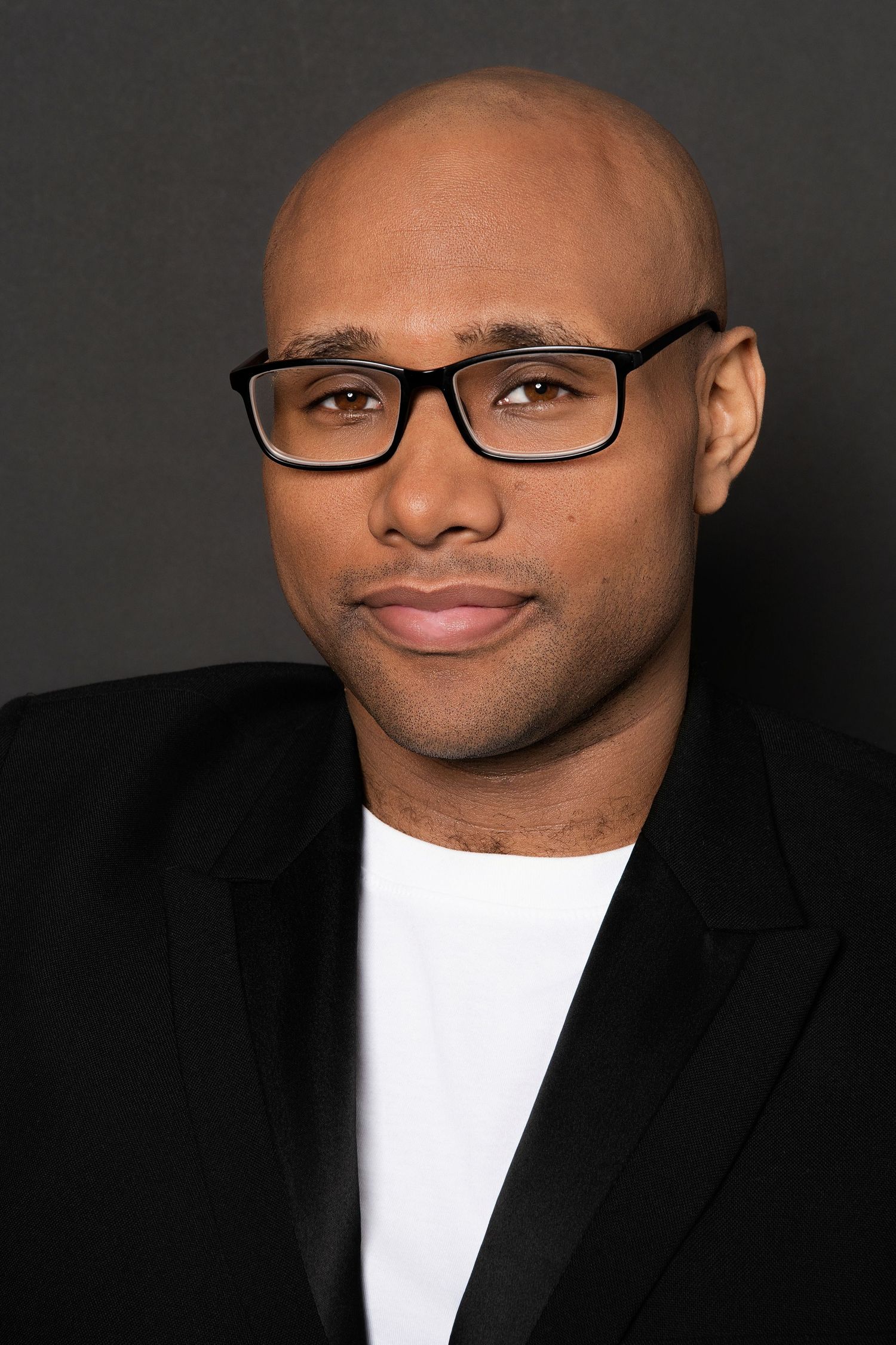 Bald Black man wearing black blazer and white shirt with glasses posing for professional headshot.