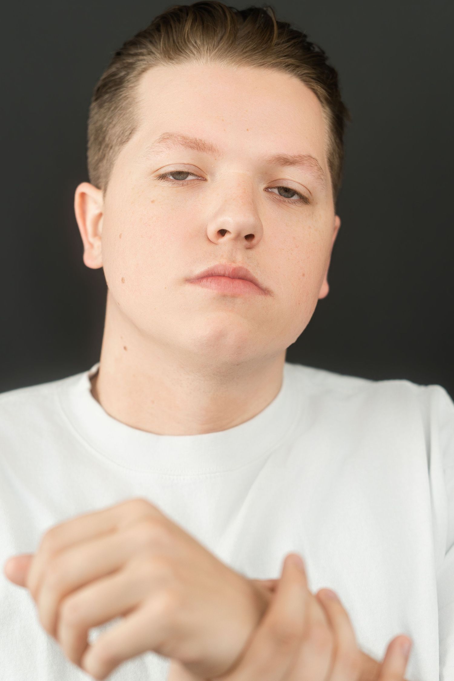 Young man in white t-shirt clasping hands, neutral expression against dark background.