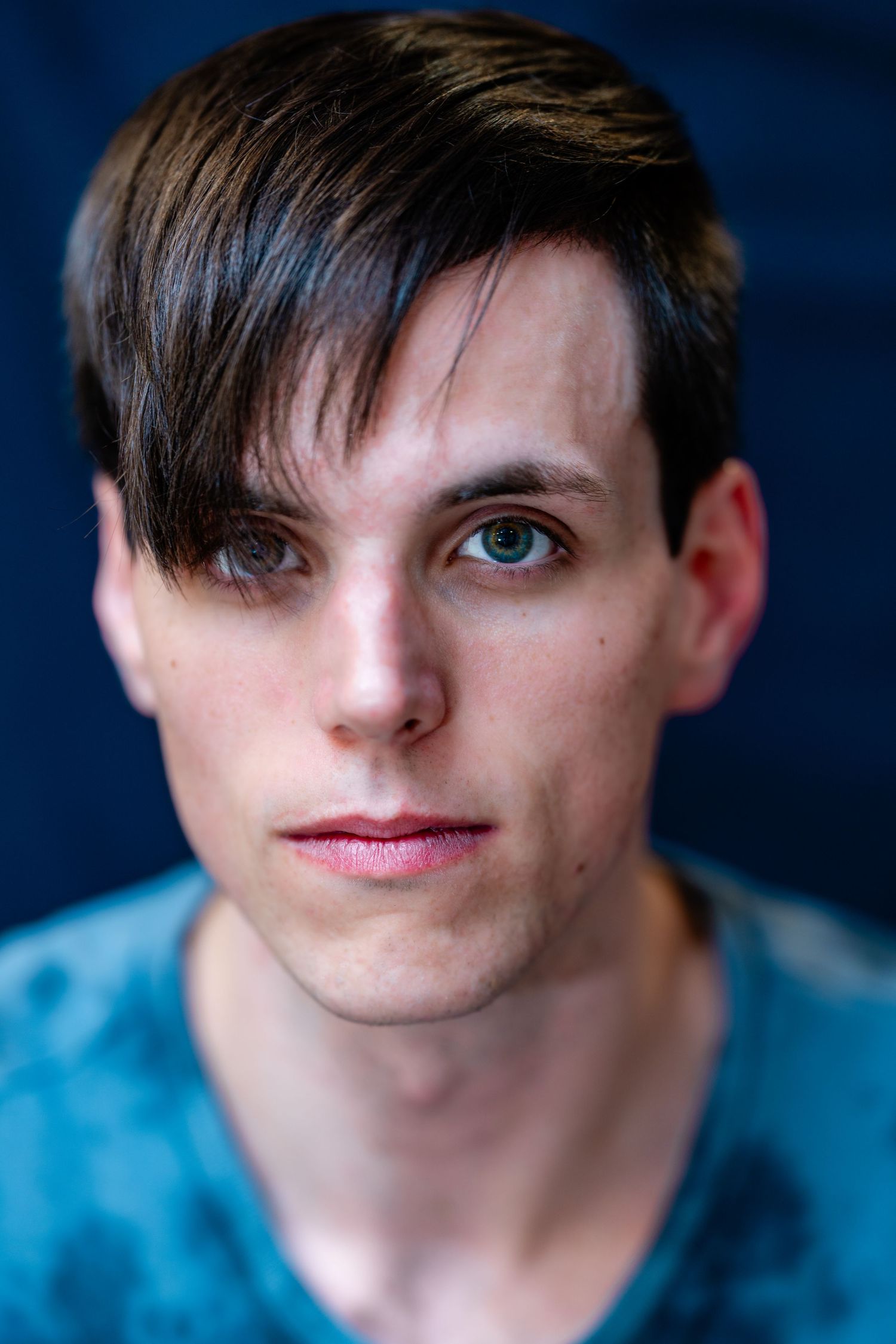 Young man with dark side-swept hair and light eyes in blue shirt, blue background.