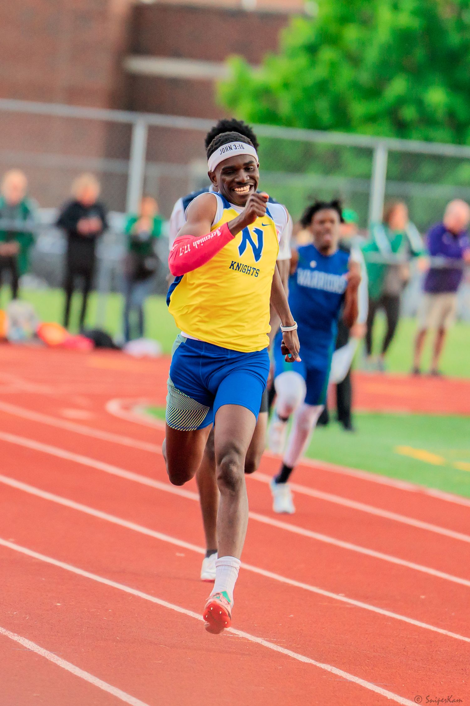 Male Knights sprinter in yellow uniform smiles while racing on a red outdoor track.