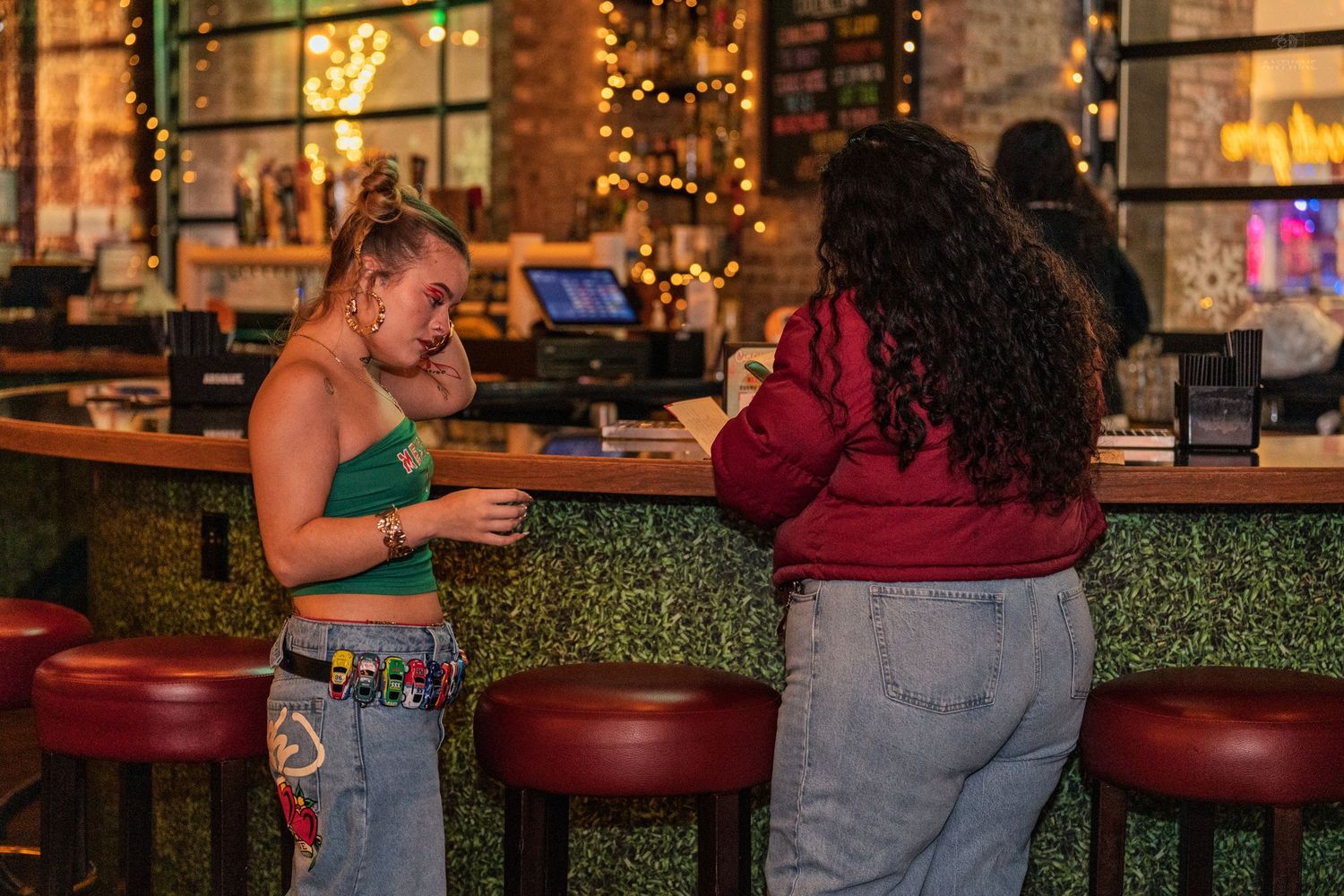 Two women conversing and sharing drinks at a green-tiled bar with warm ambient lighting.