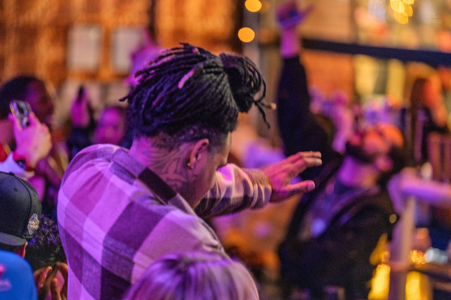 Man with dreadlocks and tattoos gestures at a lively nightclub event under purple lights.