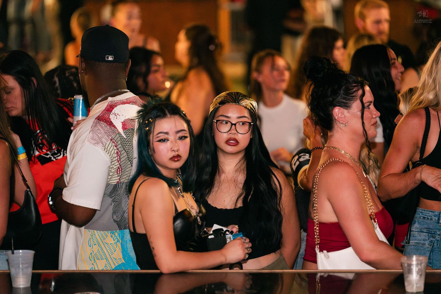 Young women socializing at a crowded nightclub bar, one wearing glasses, drinks visible on counter.