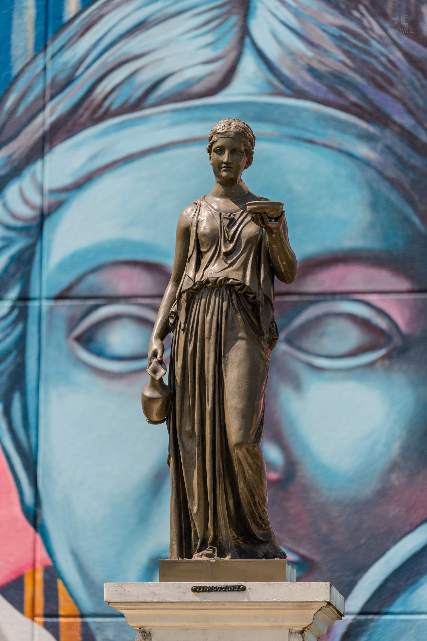 Bronze classical female statue on pedestal before vibrant blue mural with face, blending ancient art with modern street art.