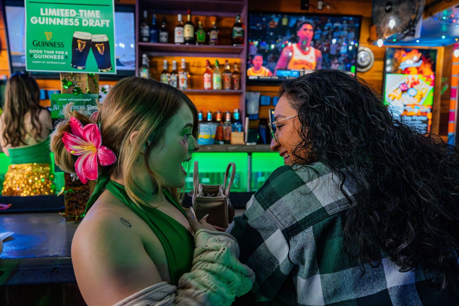 Two women laughing together at a bar with green lighting and a Guinness draft sign in the background.