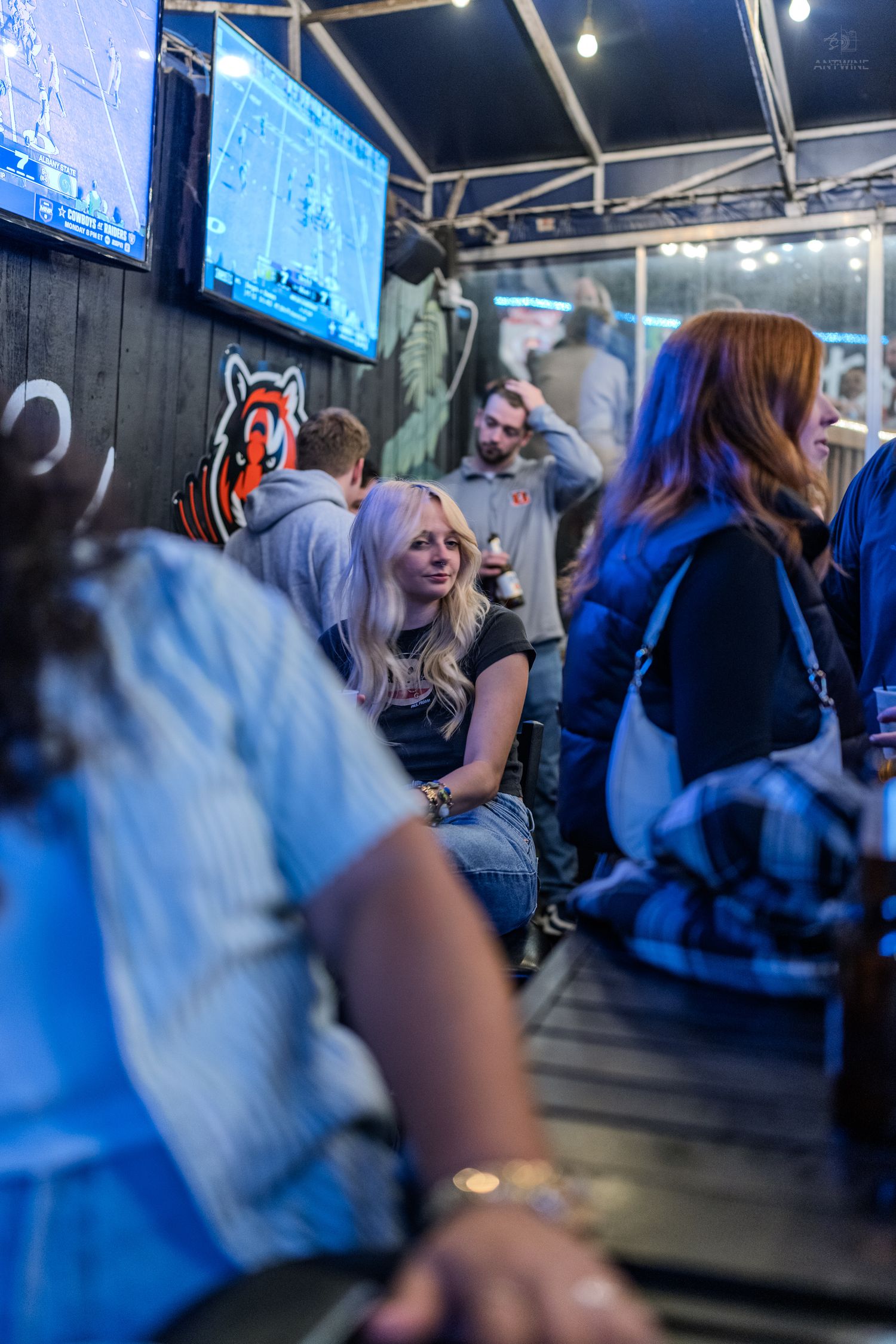 Group of people socializing at a bar with Cincinnati Bengals logo on the wall and TV screens.