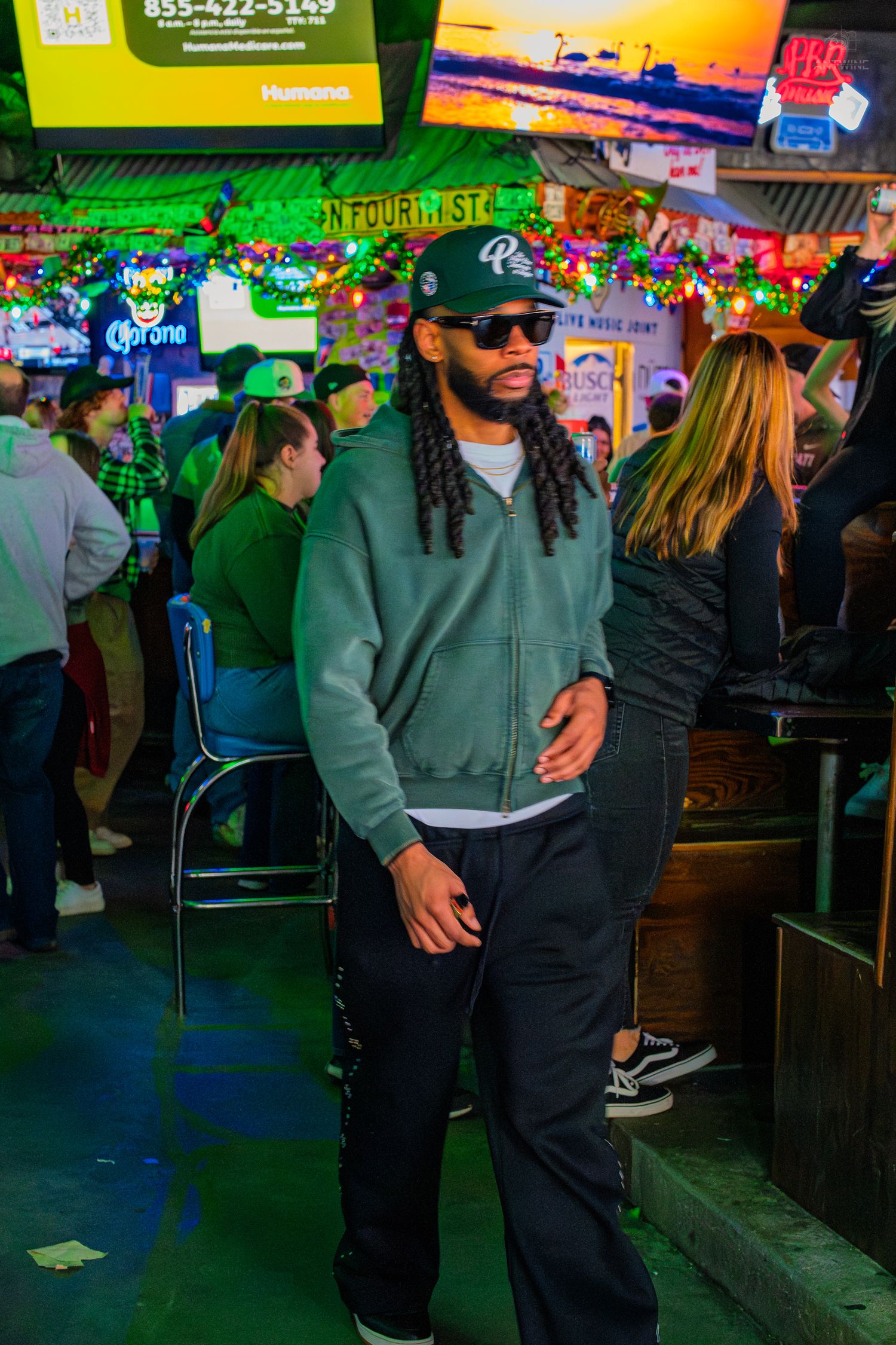 Man with dreadlocks in gray hoodie and sunglasses stands in a festively decorated bar.