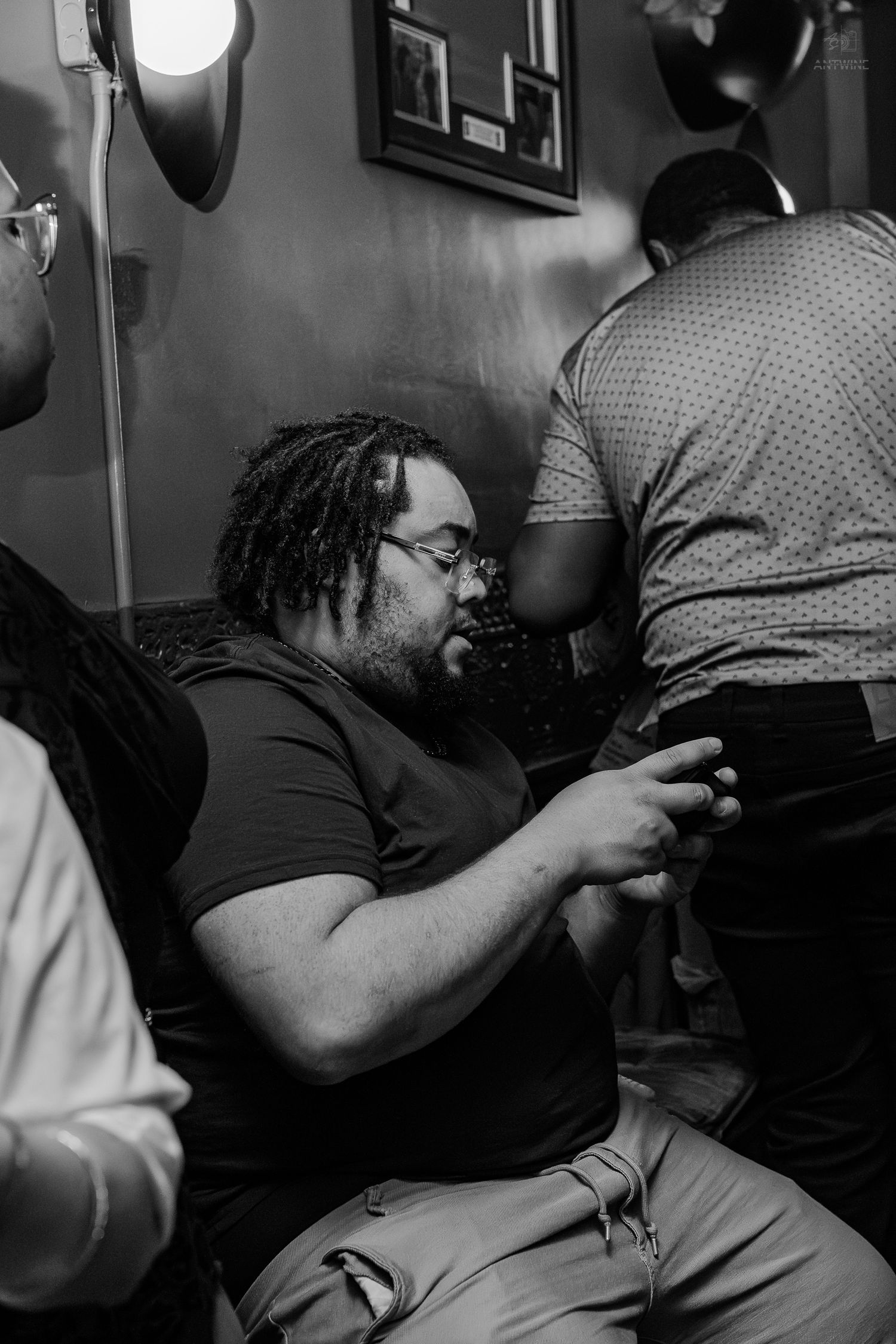 Black and white photo of a man with dreadlocks and glasses sitting and looking at his phone indoors.