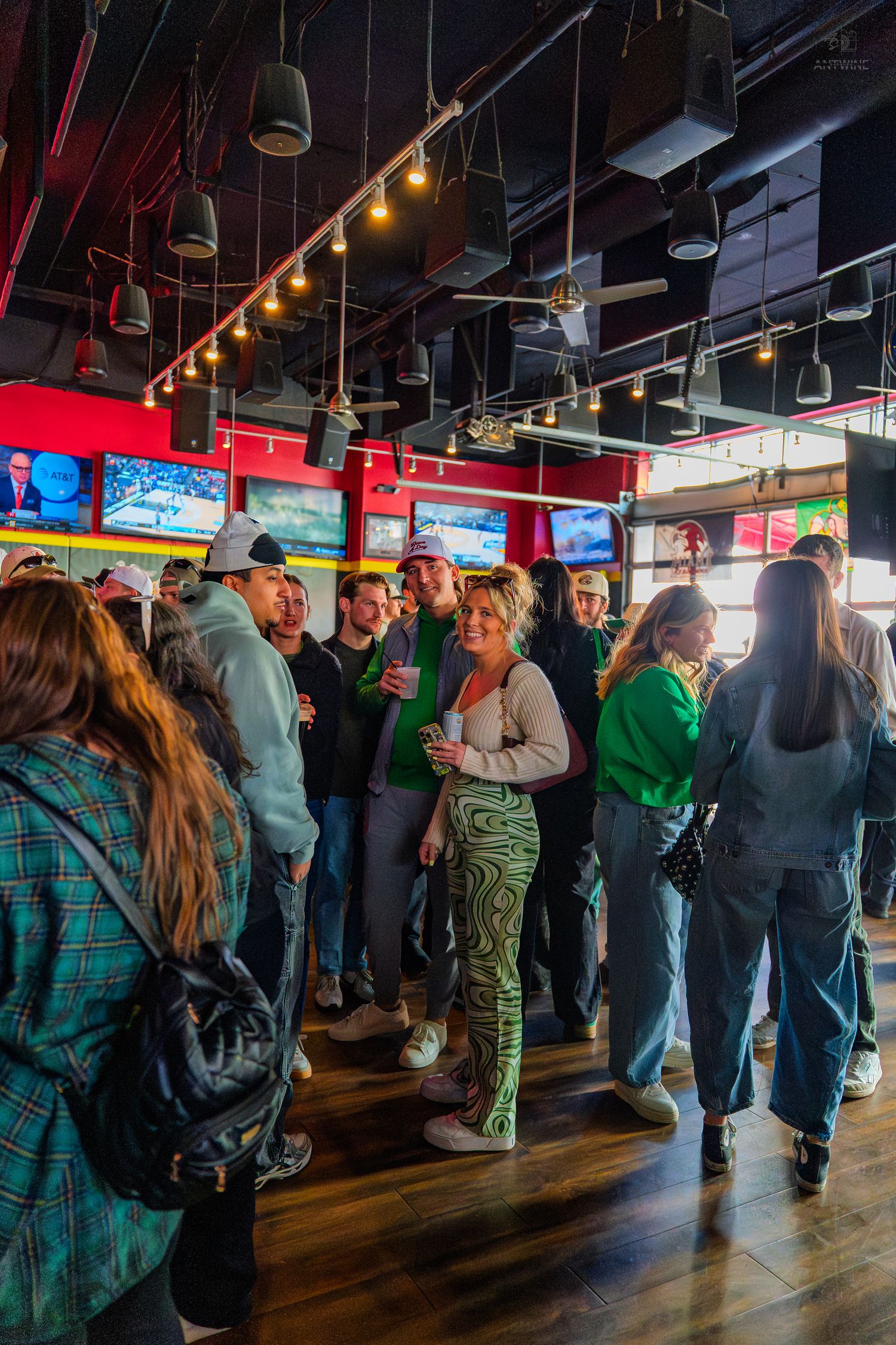 Crowd of people wearing green celebrating St. Patrick's Day at a sports bar with TVs and wood floors.