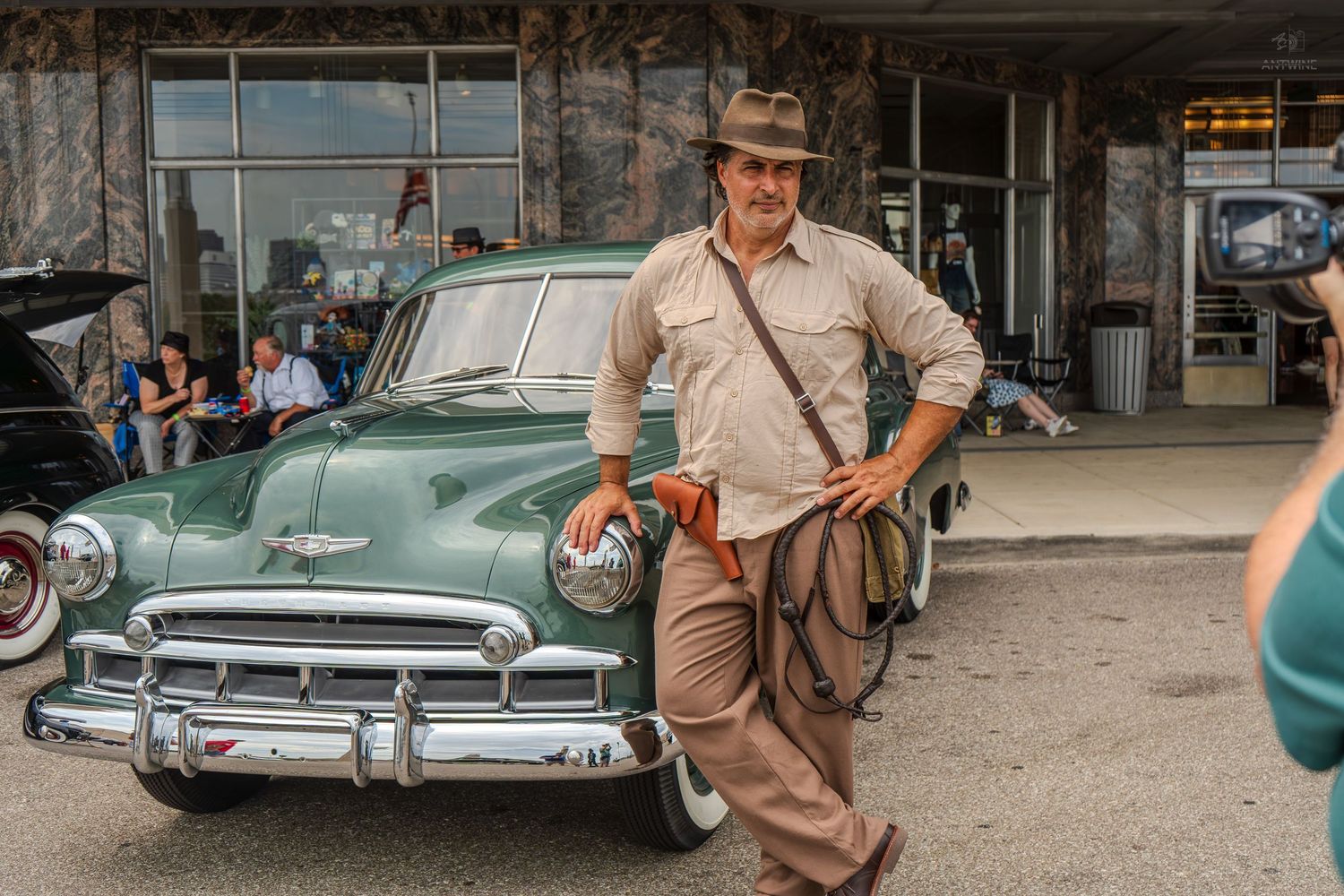 Man dressed as Indiana Jones leaning on a vintage green Chevrolet car at an outdoor classic car show.