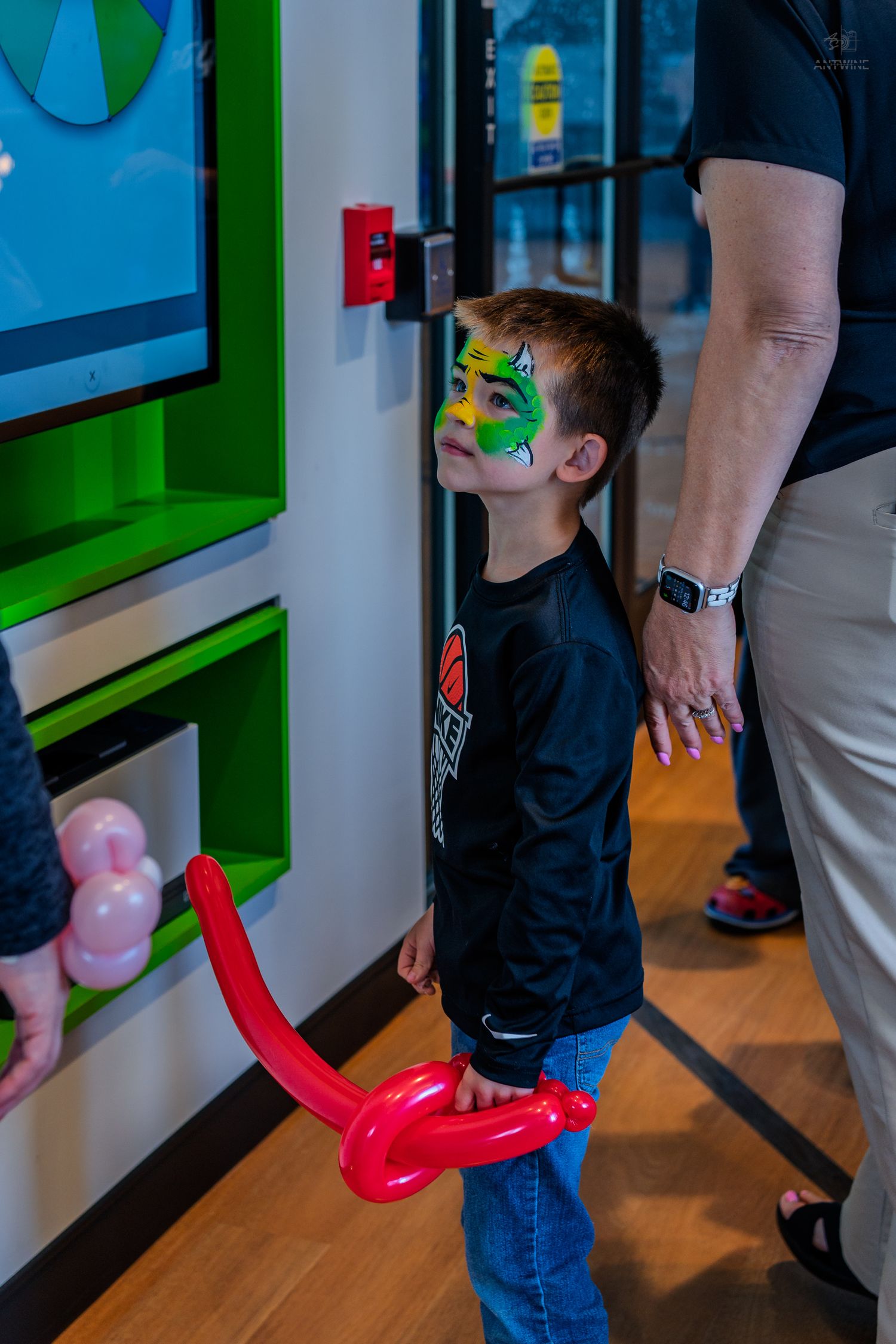 Young boy with colorful face paint holds a red balloon animal while waiting in line at an indoor event.