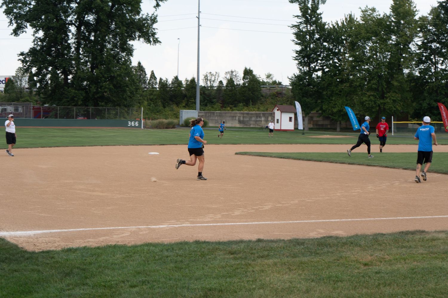 Players compete in a recreational softball game on a dirt infield diamond surrounded by green trees on a sunny day.