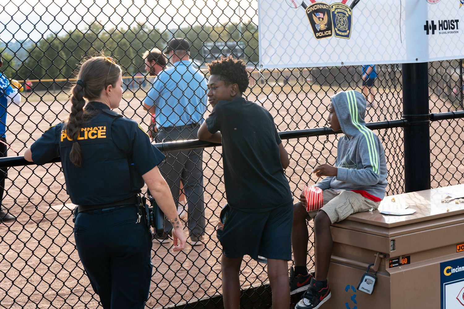 Female police officer speaks with two children near a chain-link fence at an outdoor community event.
