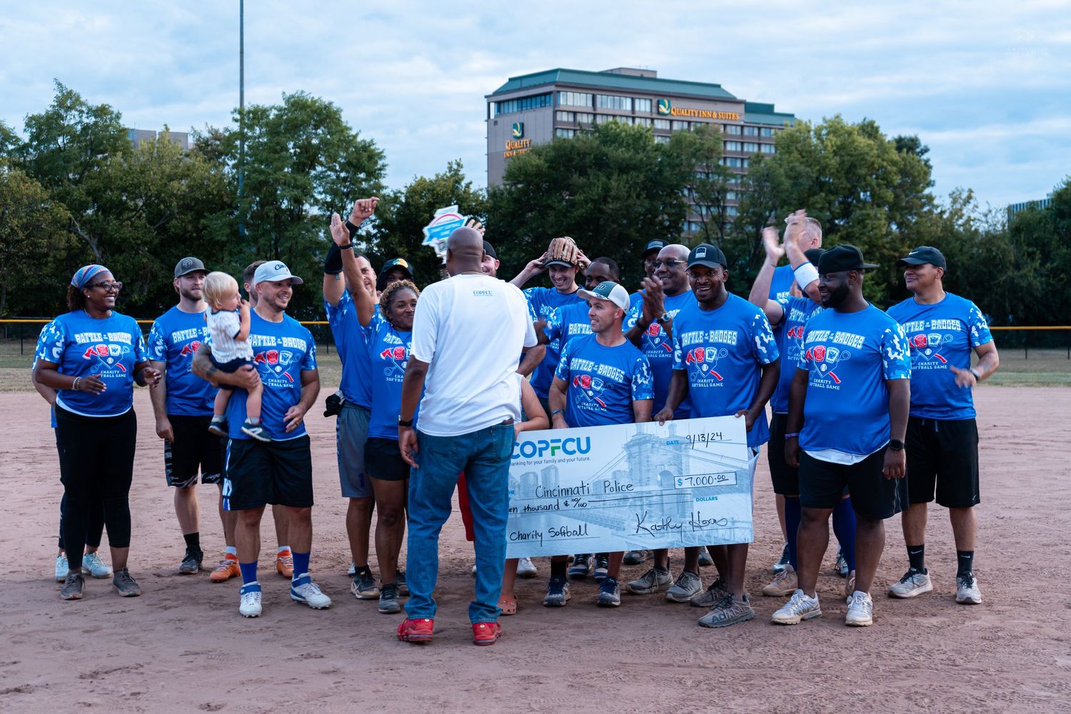 Softball team in blue COPFCU shirts celebrate with a large ceremonial check on an outdoor baseball diamond.