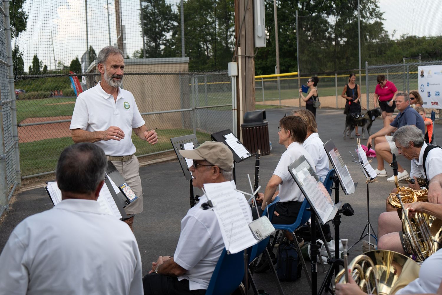 Conductor leads an outdoor brass band performance at a baseball field on a sunny day.