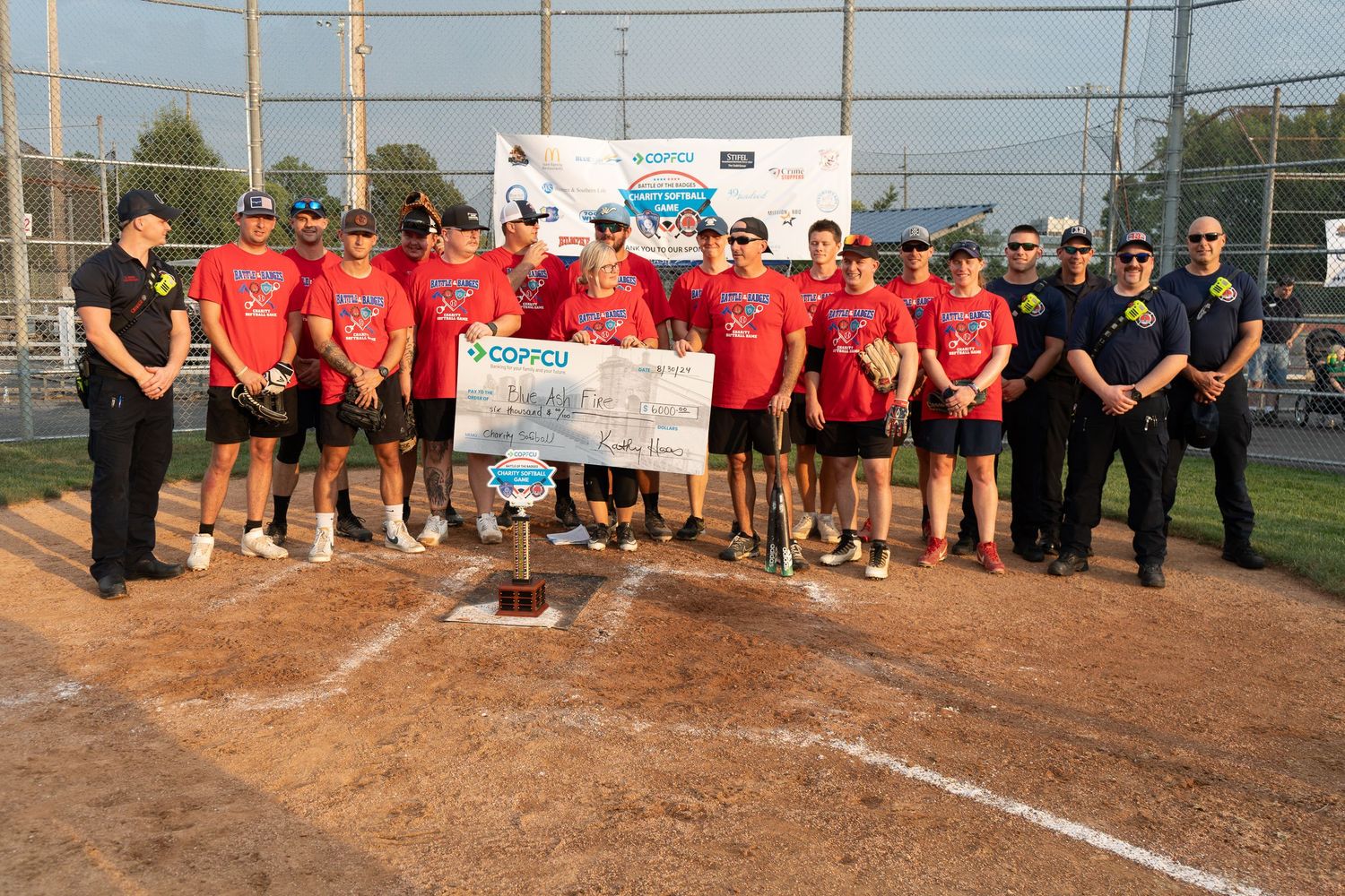 Team in red jerseys poses with oversized COPCU check and trophy on softball field with officials.