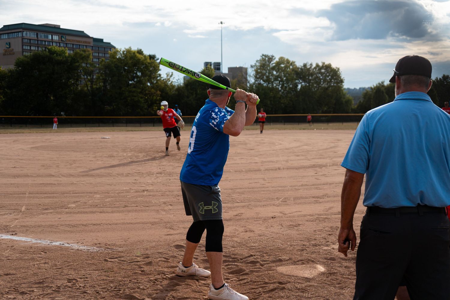 Softball batter in blue jersey swings a green bat during an evening recreational league game with umpire nearby.