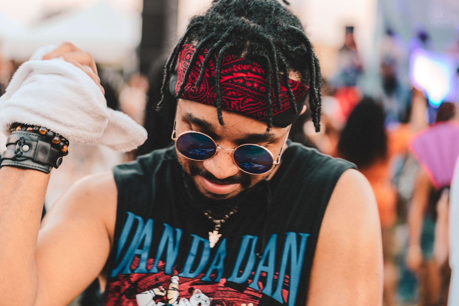 Young man with red-wrapped dreadlocks and round sunglasses at an outdoor music festival.