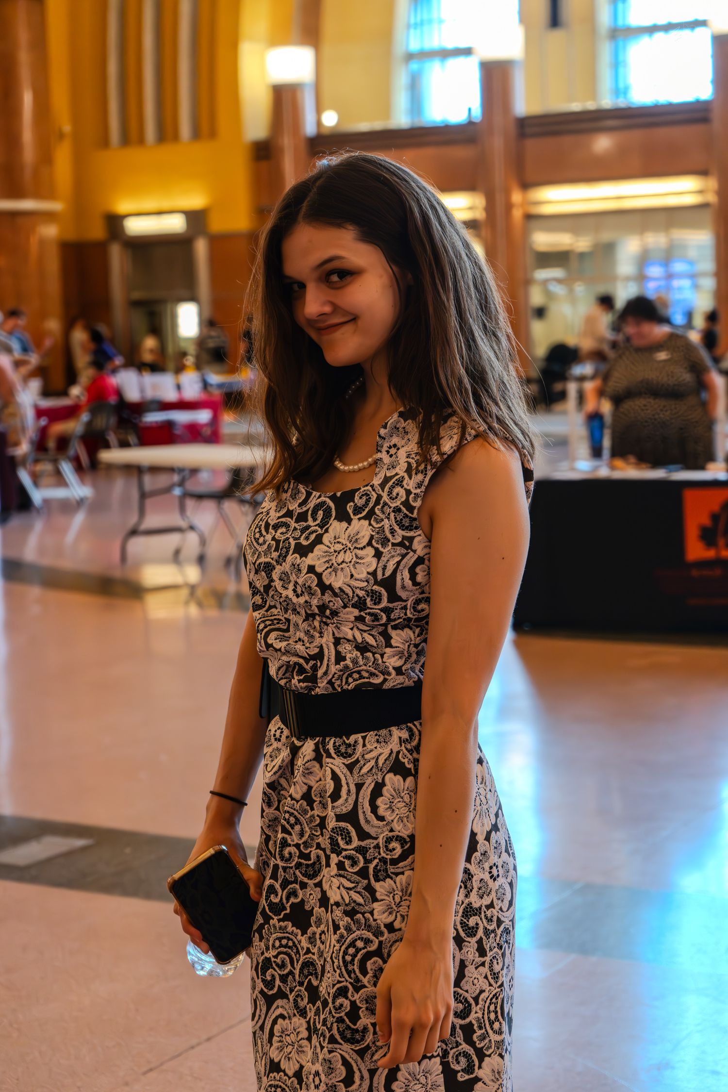 Young woman in black and white floral dress smiling in a large indoor hall.