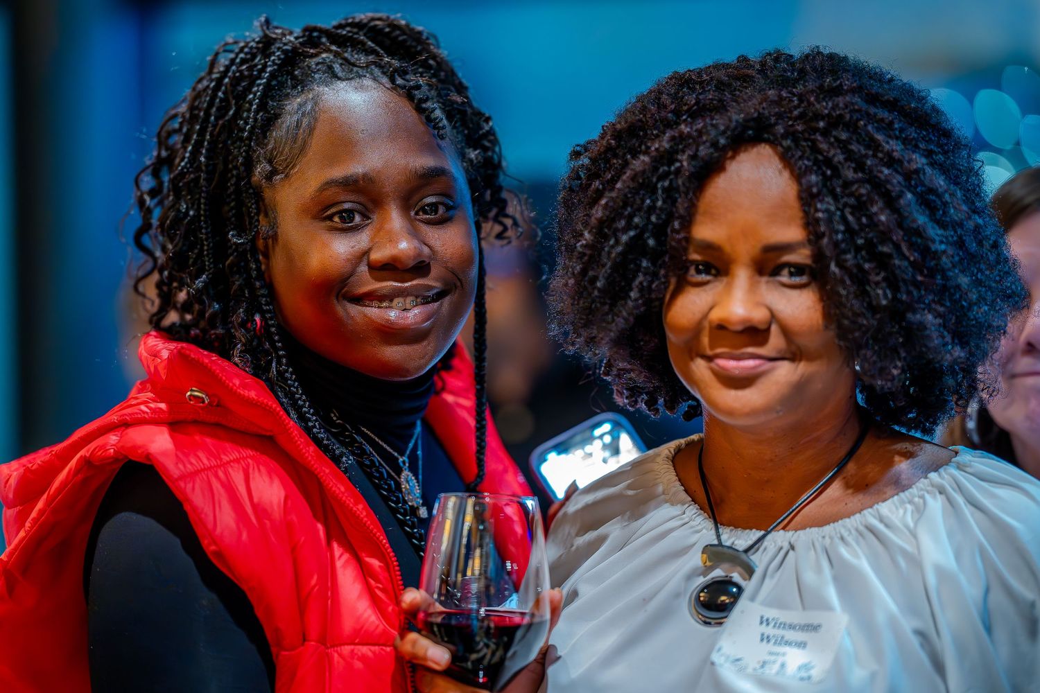 Two women smiling at an indoor event, one in a red vest holding a wine glass, posing together for a photo.