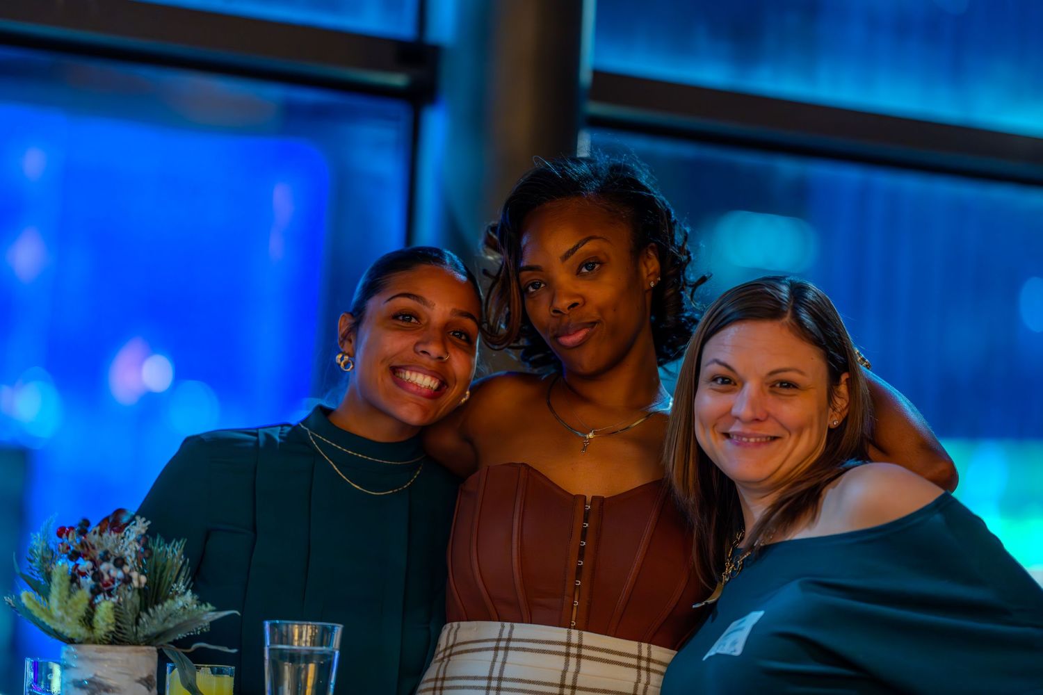 Three women smiling and posing together at an evening event with blue ambient lighting in the background.